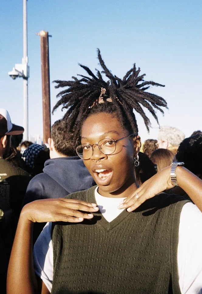 Photo of Irawo Ajasin, a Black nonbinary individual with glasses, posing with their hands around their face outside in a crowd of people.