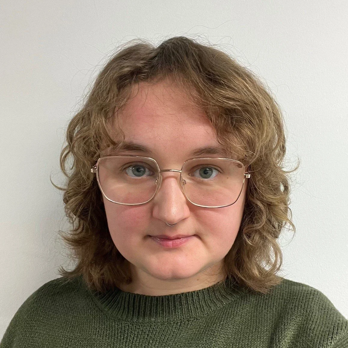A photo of Megan Fereday standing against a white wall. They are a white nonbinary person, with square thin glasses, a textured olive green sweater with mushrooms on it, and layered blond curls.