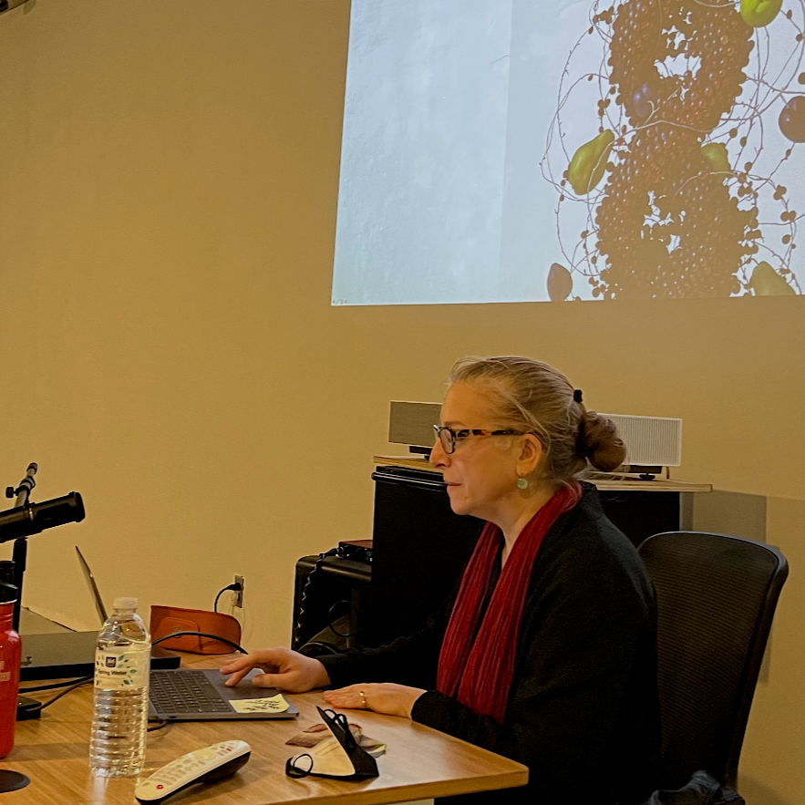 A photo of Mia Brownell giving a talk, sat at a desk with a microphone, a projected image behind her