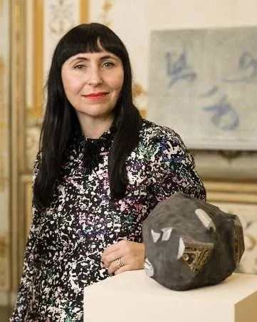 Agnieszka Kurant has dark straight hair, a colorful blouse, and poses beside a sculpture
