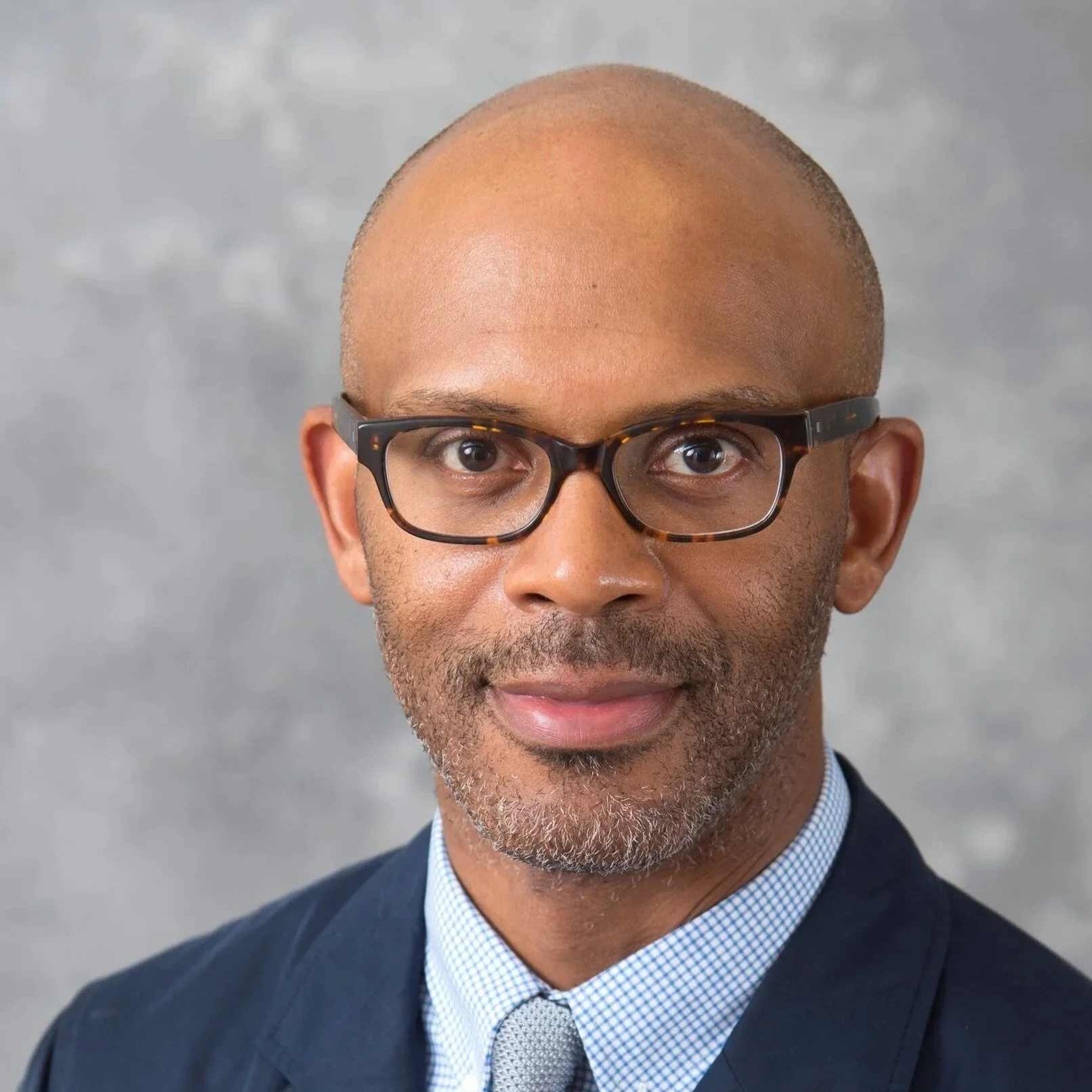 A photo of Rayvon Fouche, a Black man with glasses and a blue suit against a gray background