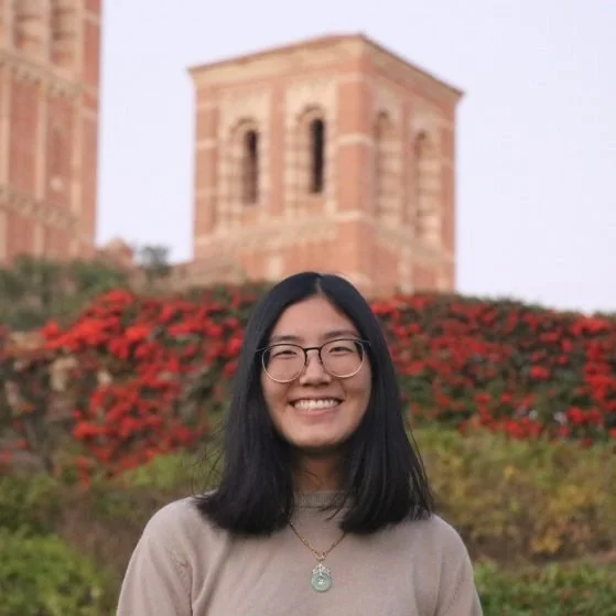 Portrait of Cherice Chan, an Asian woman with glasses, smiling in front of flowers and a brick building