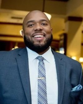 A photo of Aaron Dial, a Black man with dark facial hair, a blue suit, and a smile