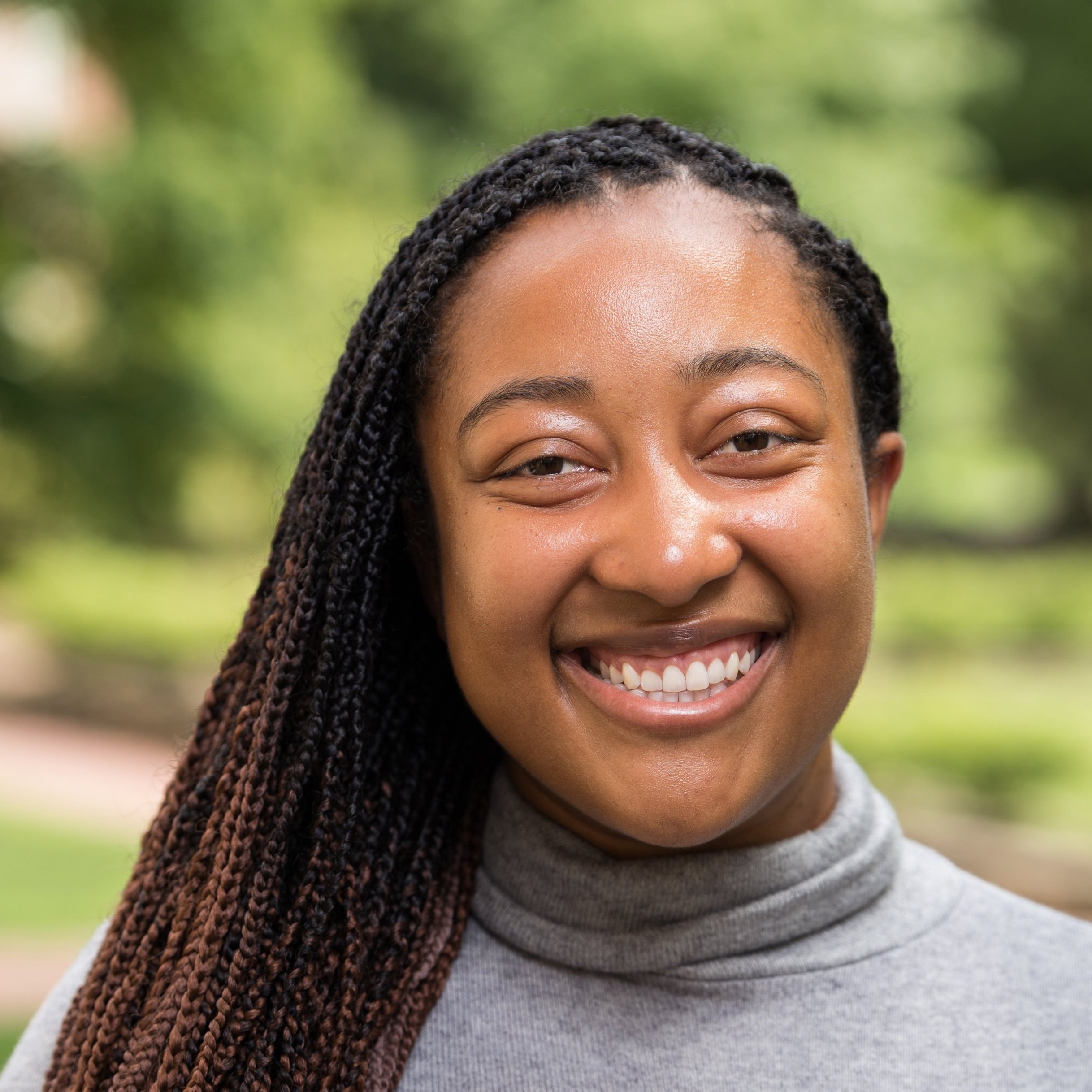 A photo of LaRisa, a Black woman with ombre braids and a light gray turtleneck sweater. Standing outside, the background of the photo is out of focus greenery.