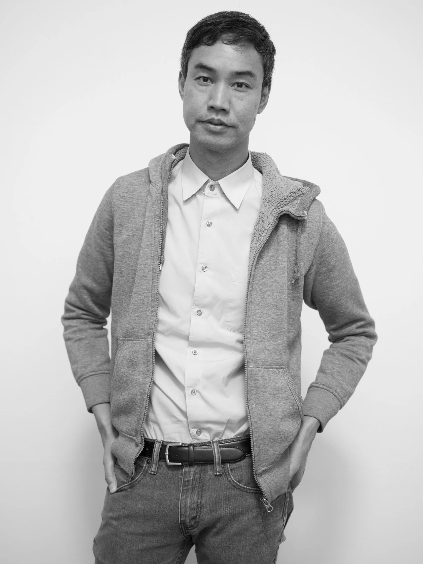 Miljohn is pictured in black and white, with short dark hair, a gray sweater, white button up, and jeans