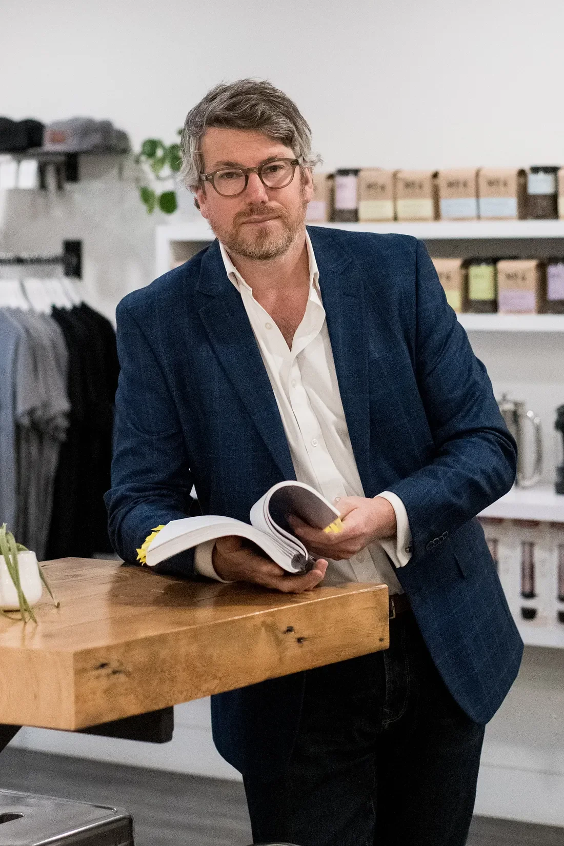 A man with glasses, wearing a navy blazer and white shirt, standing at a wooden counter in a store. He is holding an open book or catalog. Behind him are shelves with boxes and jars, and a clothing rack.