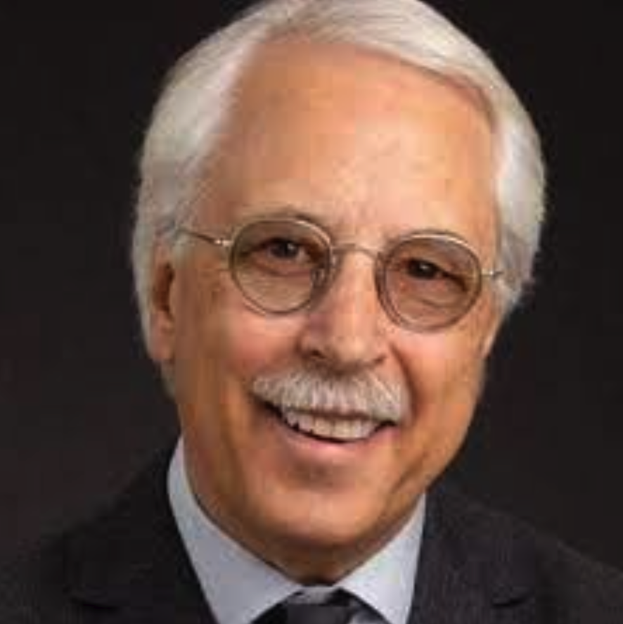 Portrait of an elderly man with white hair, glasses, and a mustache, smiling, wearing a dark suit with a white shirt and tie.