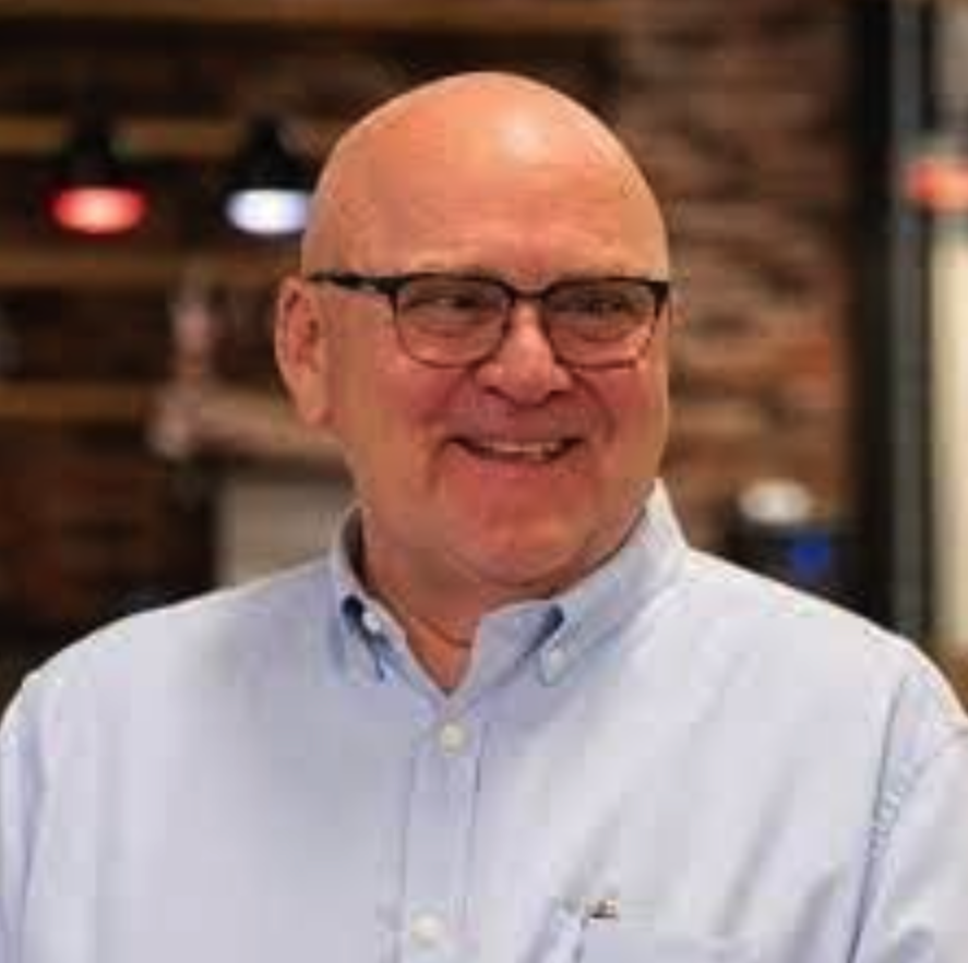 A bald man wearing glasses and a light blue collared shirt smiling in a restaurant or cafe setting with a brick wall background.