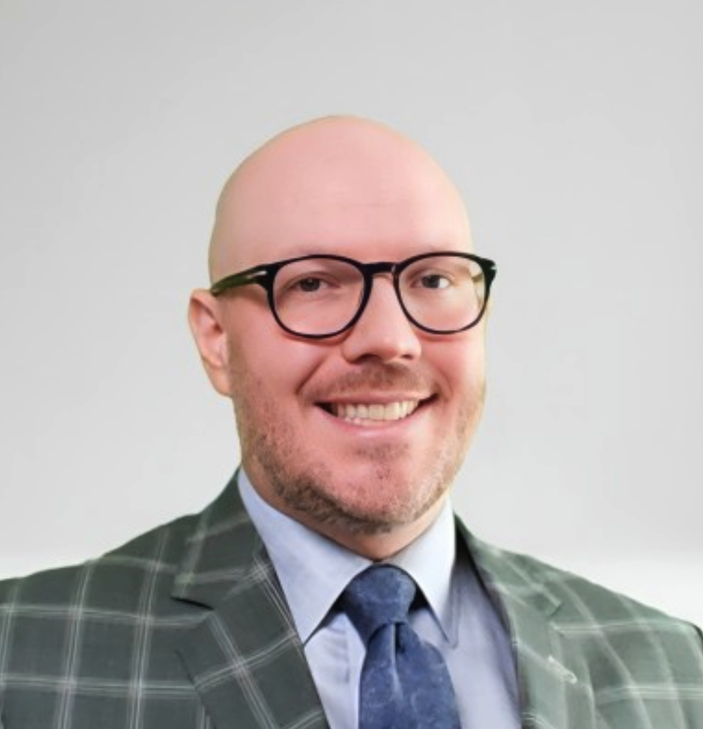 A smiling man with glasses, a light blue shirt, a blue tie, and a plaid grey blazer, standing against a white background.