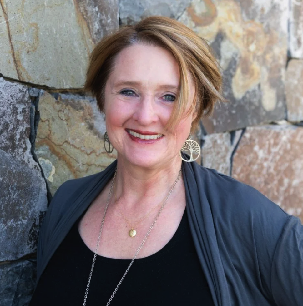 A woman with short, auburn hair smiling, wearing silver jewelry including a necklace, earrings, and a tree-of-life pendant, standing in front of a stone wall.