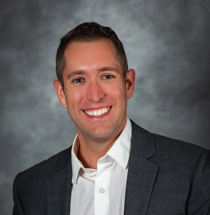 Professional headshot of a smiling man in a gray suit and white shirt against a gray background.
