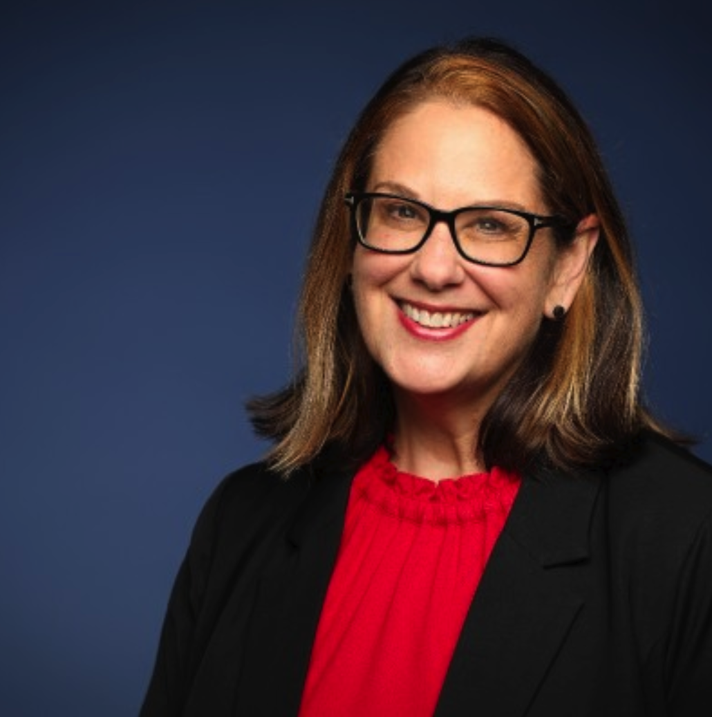 Photograph of a woman with shoulder-length brown hair, wearing glasses, a red blouse, and a black blazer, smiling against a blue background.