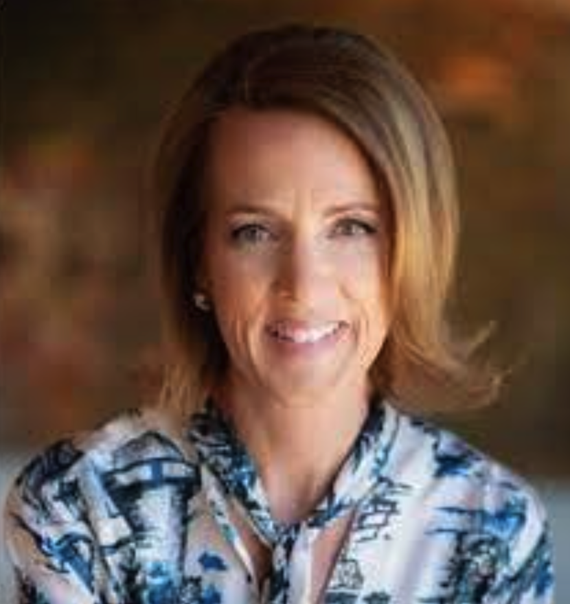 A woman with shoulder-length light brown hair smiling at the camera, wearing earrings and a colorful patterned blouse, with a blurred background.