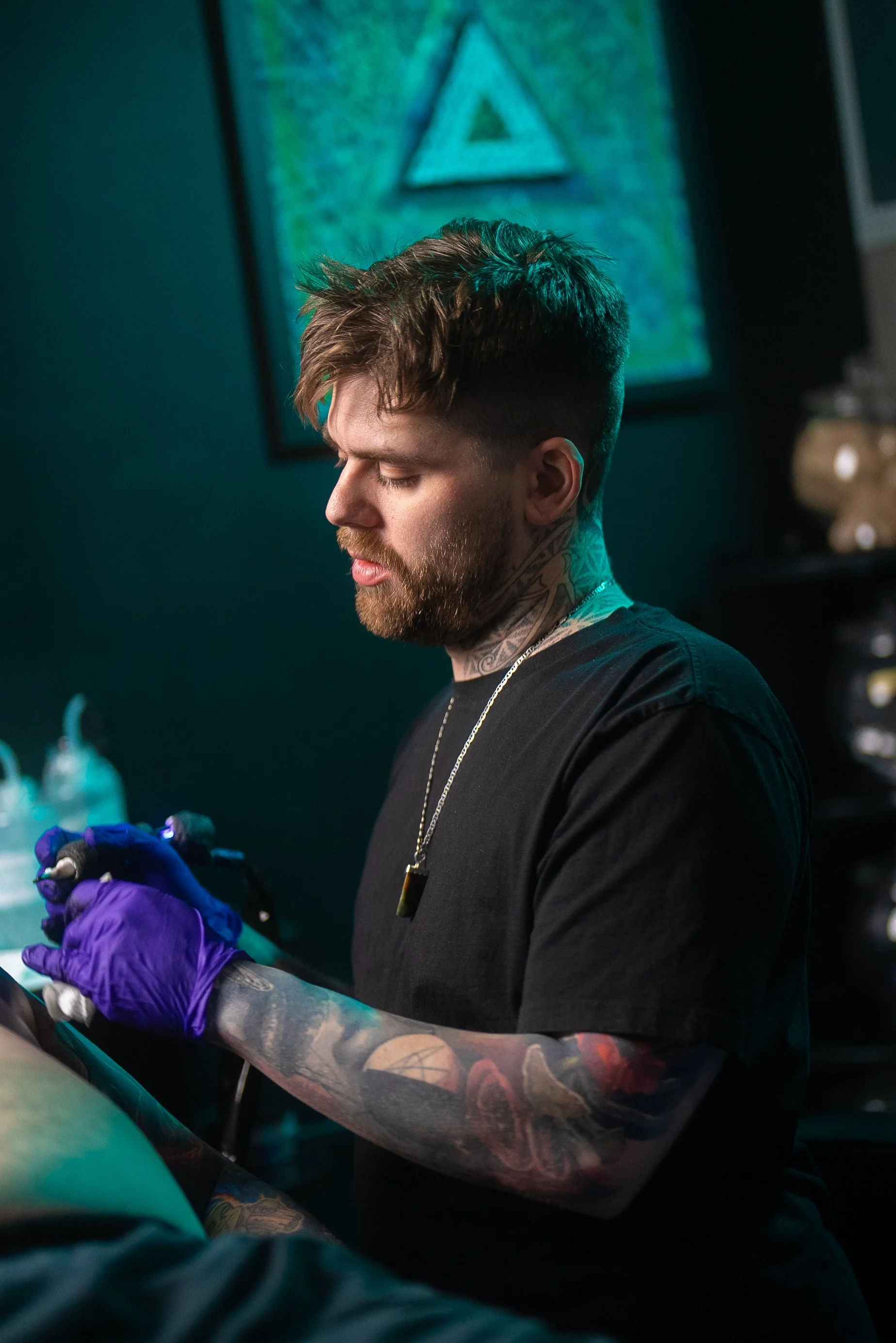 Tattooed man with brown hair and beard, wearing a black shirt and purple gloves, performing a tattoo on someone's leg in a tattoo studio