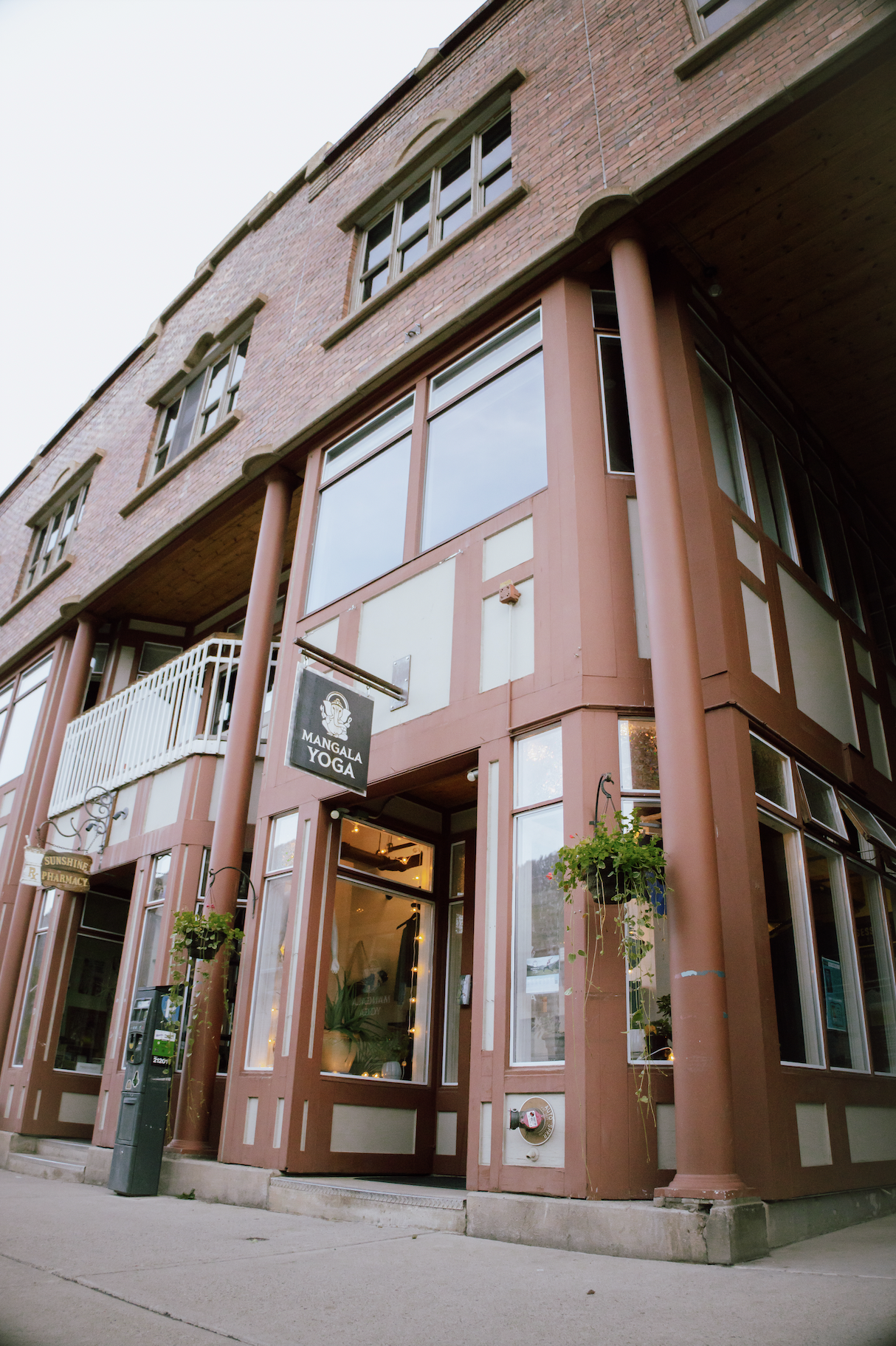 A multi-story brick building with large windows and hanging flower baskets, featuring signs for Mangala Yoga and Sunshine Pharmacy.