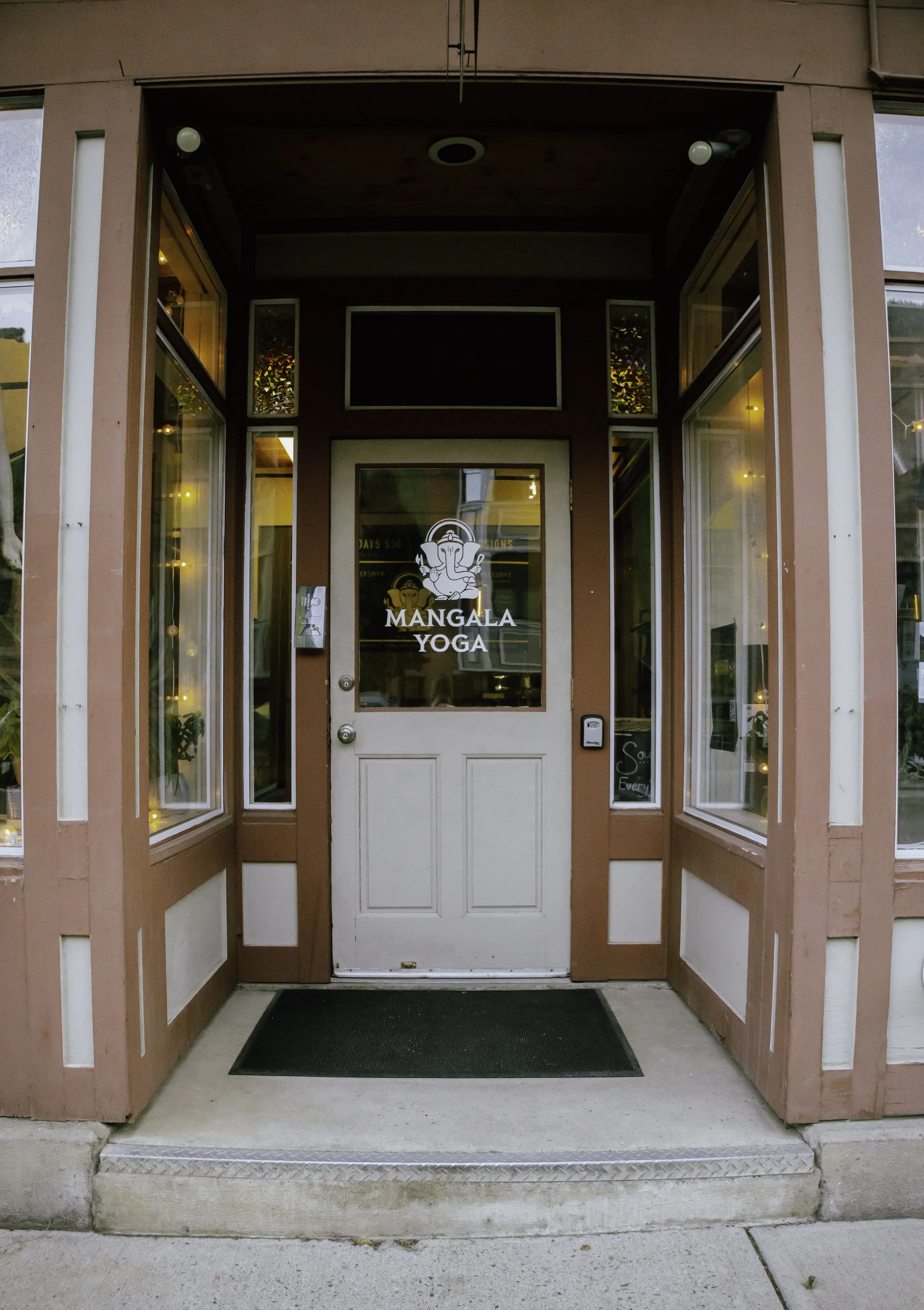 Front entrance to Mangala Yoga studio with a glass door, sign with an image of Ganesha, and side windows decorated with string lights.