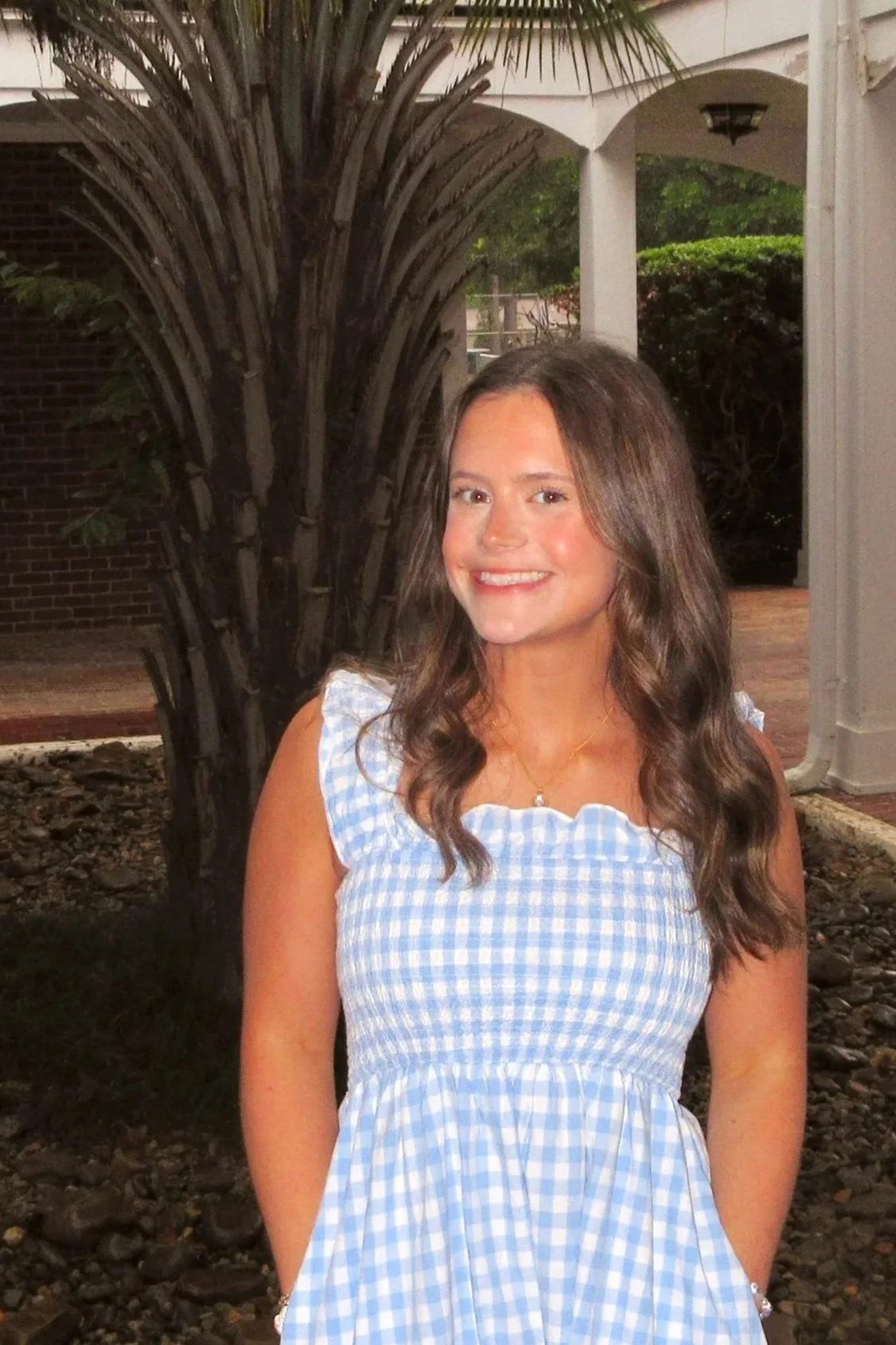 A young woman with long dark hair smiling on a white sandy beach during sunset, wearing a green and white striped button-up shirt and light blue jeans.