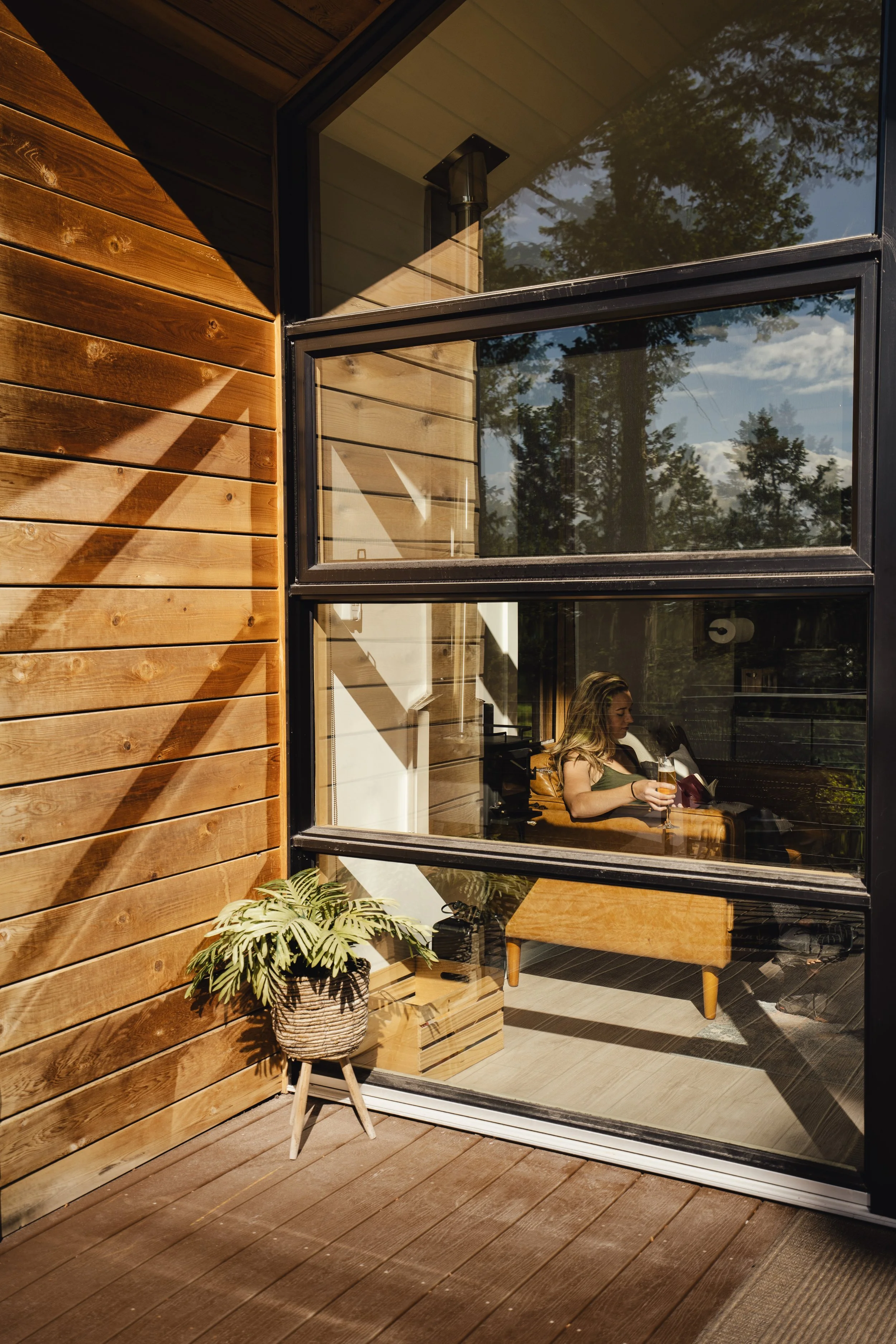 A person sitting by a window in a room with wooden walls, holding a drink, with a potted plant outside on a wooden deck.