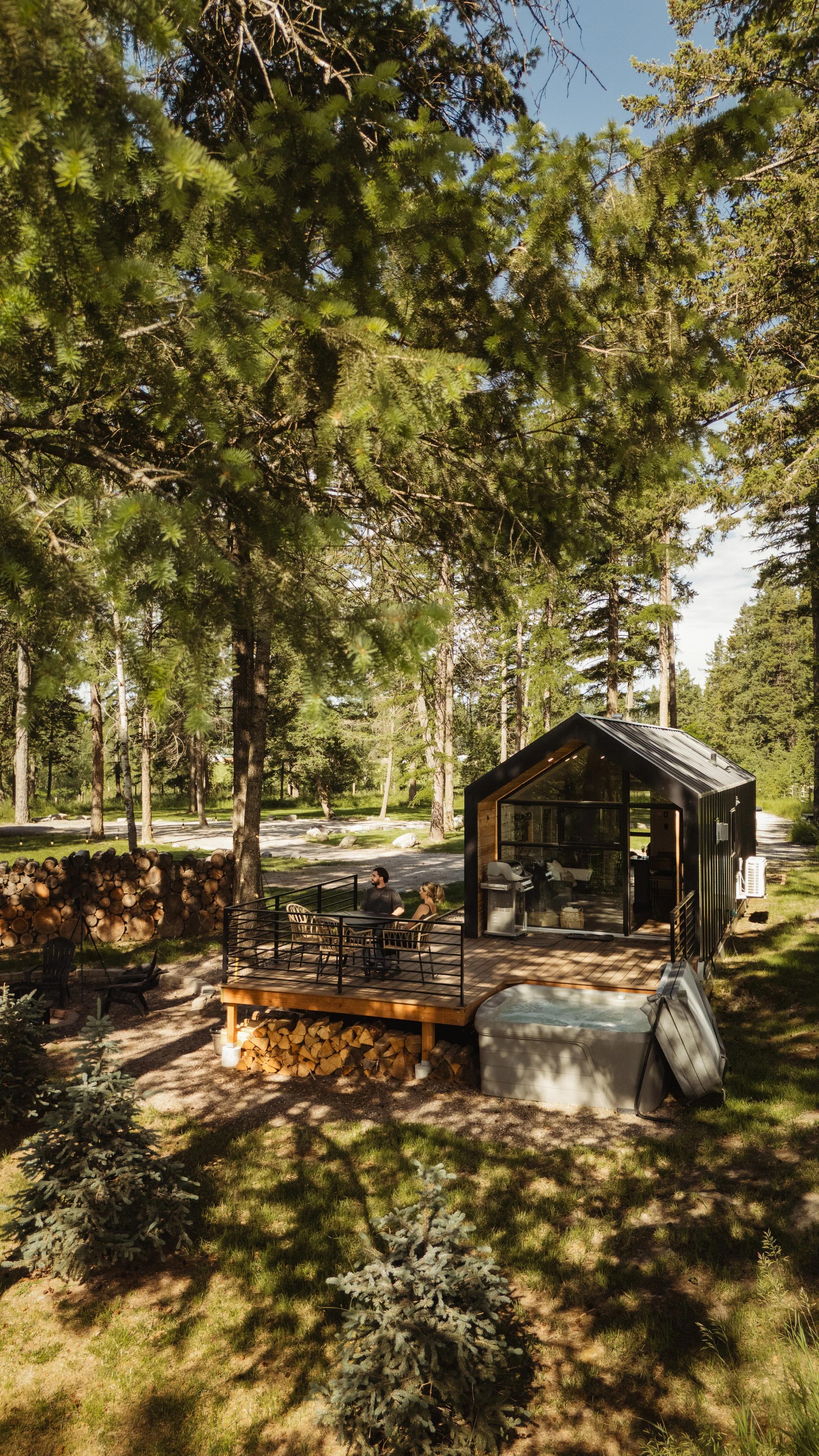 A wooded backyard with tall trees and a modern black shed. Two people are sitting at a table on a deck, enjoying an outdoor setting, with wood logs stacked nearby.
