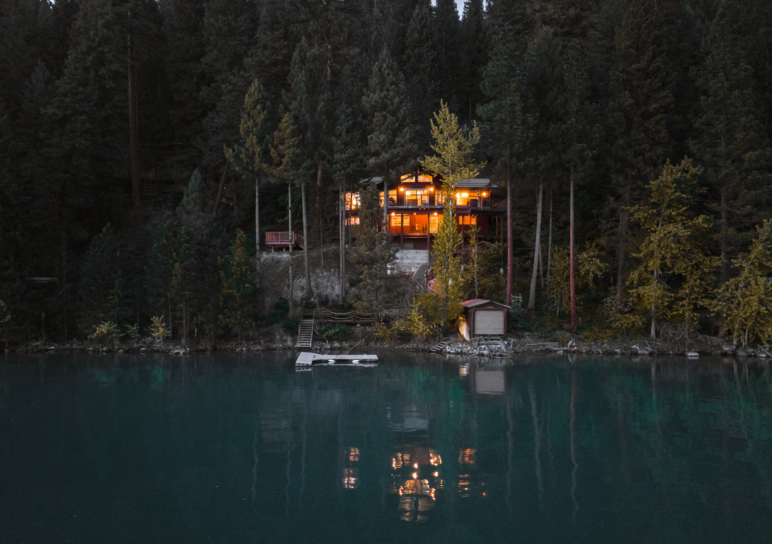 A house with large windows illuminated from within is nestled among tall trees on a hillside, overlooking a calm body of water at dusk.
