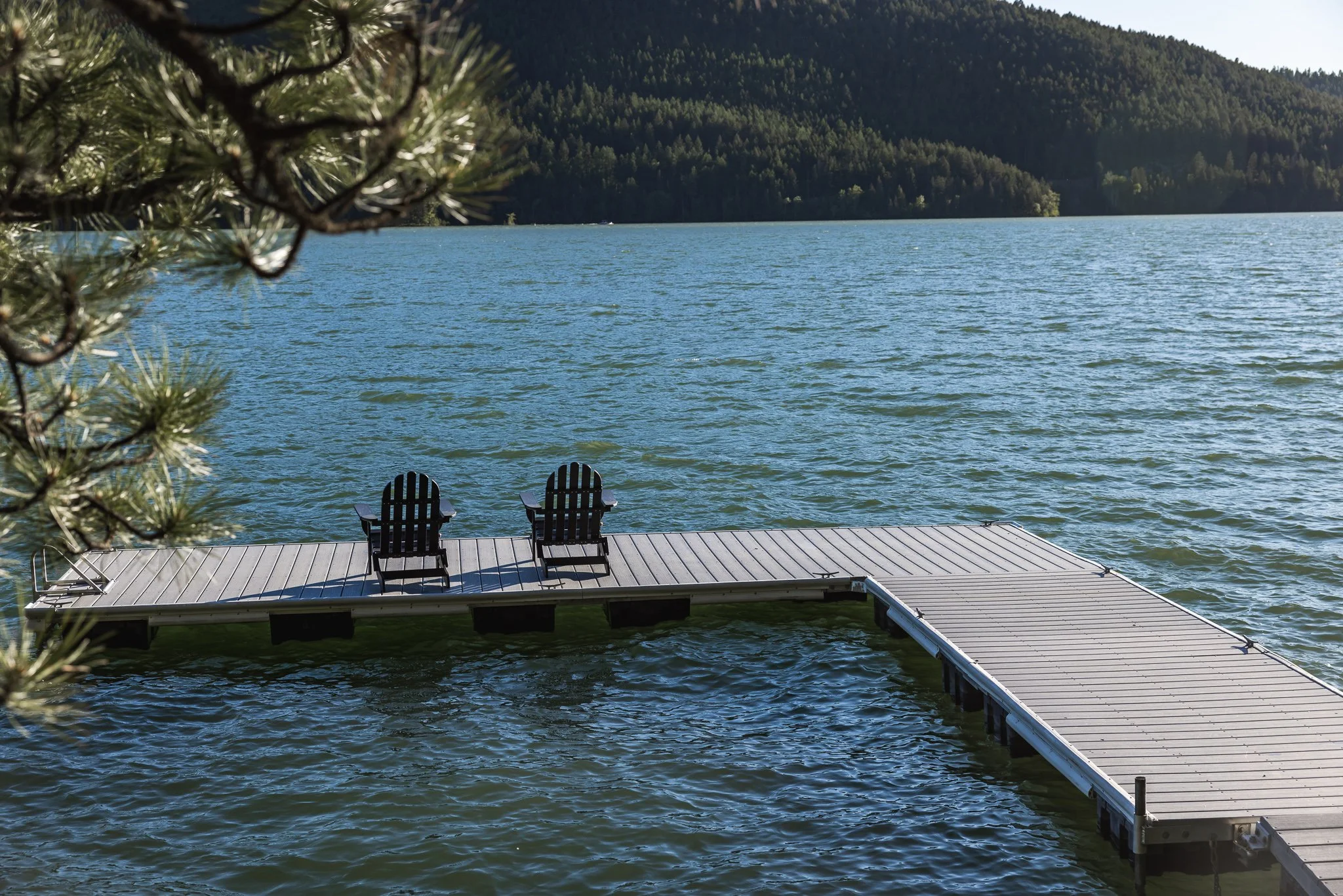 A lakeside scene with two wooden chairs on a dock, overlooking a lake with mountains in the background, and a tree branch partially visible on the left side.