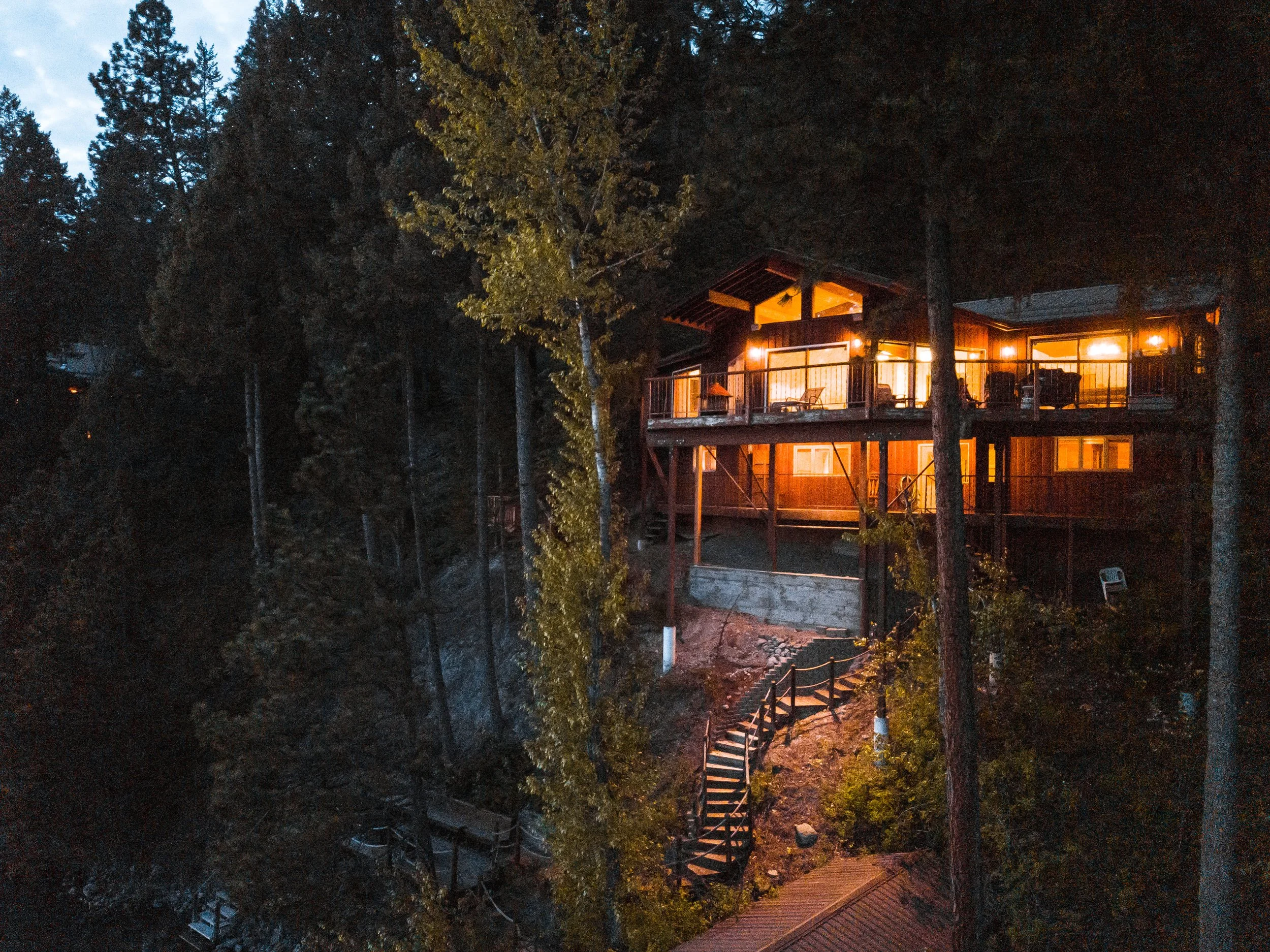 A multi-story house illuminated with warm yellow lights, nestled among tall trees at dusk or nighttime, with a staircase leading down to a lower level.