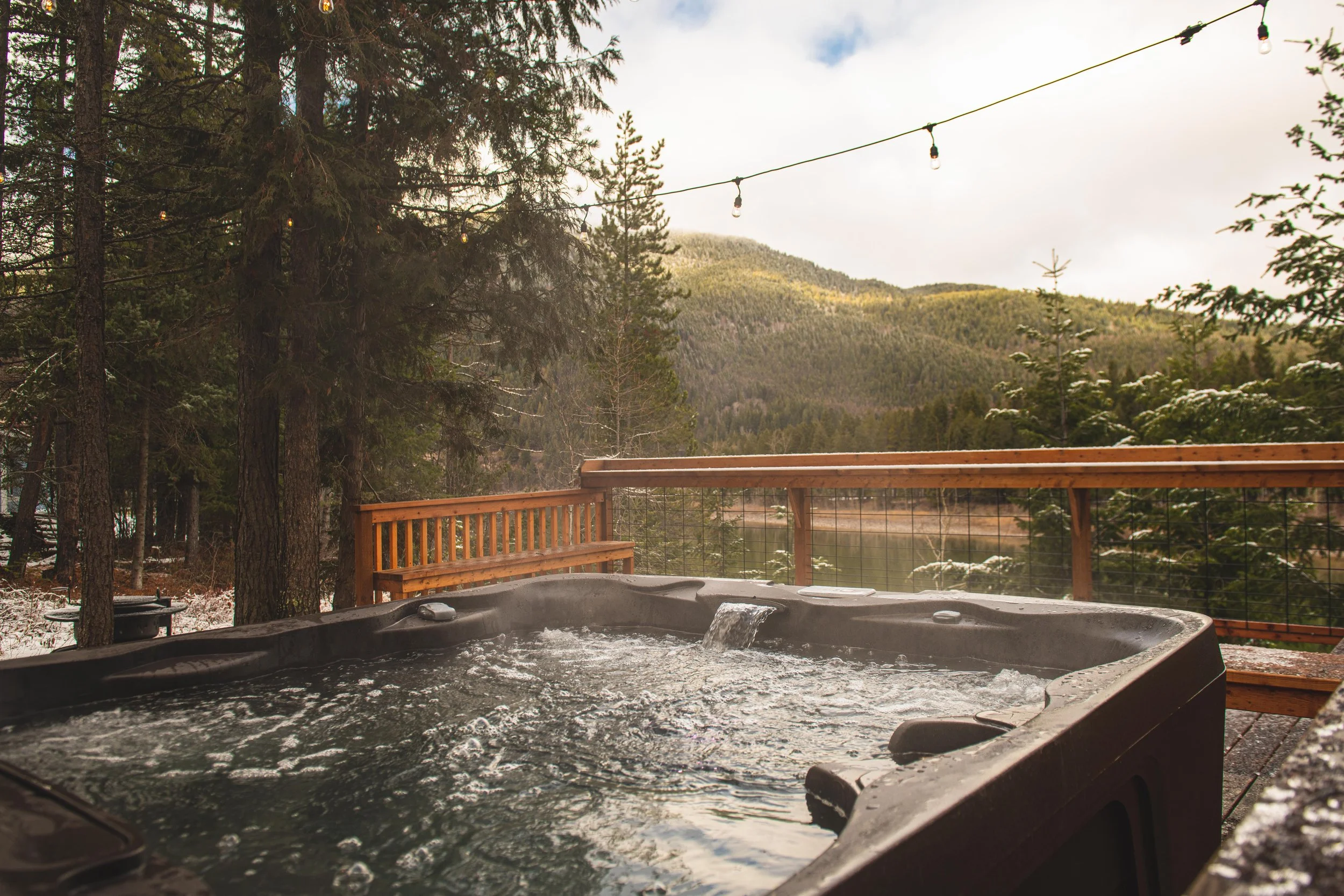An outdoor hot tub on a wooden deck, overlooking a scenic mountain landscape with pine trees and a cloudy sky. String lights are hanging above the deck.