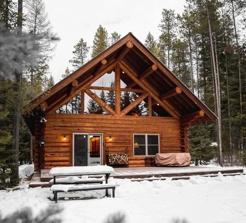 A wooden cabin in a snowy forest, with a deck, outdoor furniture, and string lights.