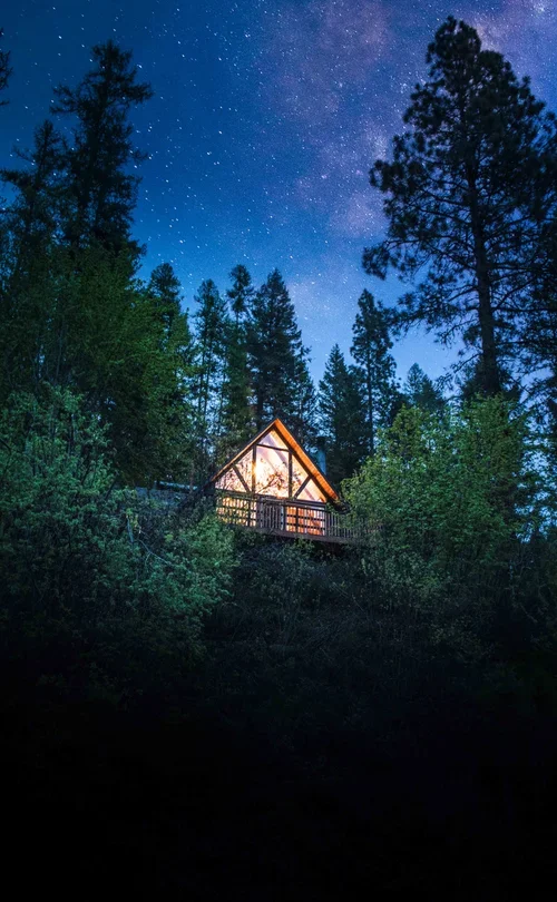 A small A-frame cabin illuminated inside, situated on a wooded hillside under a starry night sky.