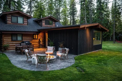 Exterior view of a modern house with a wooden and dark siding, featuring a backyard patio with outdoor furniture and a fire pit, surrounded by trees.