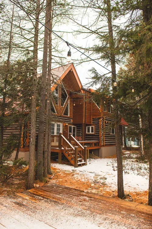 A wooden cabin in a forest with pine trees, a snowy ground, and a dirt road leading to the stairs.