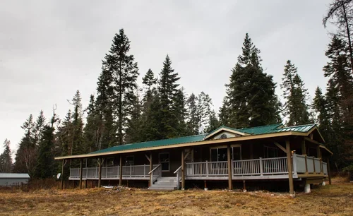 A large house or cabin with a long covered porch, situated in a wooded area with tall pine trees in the background.