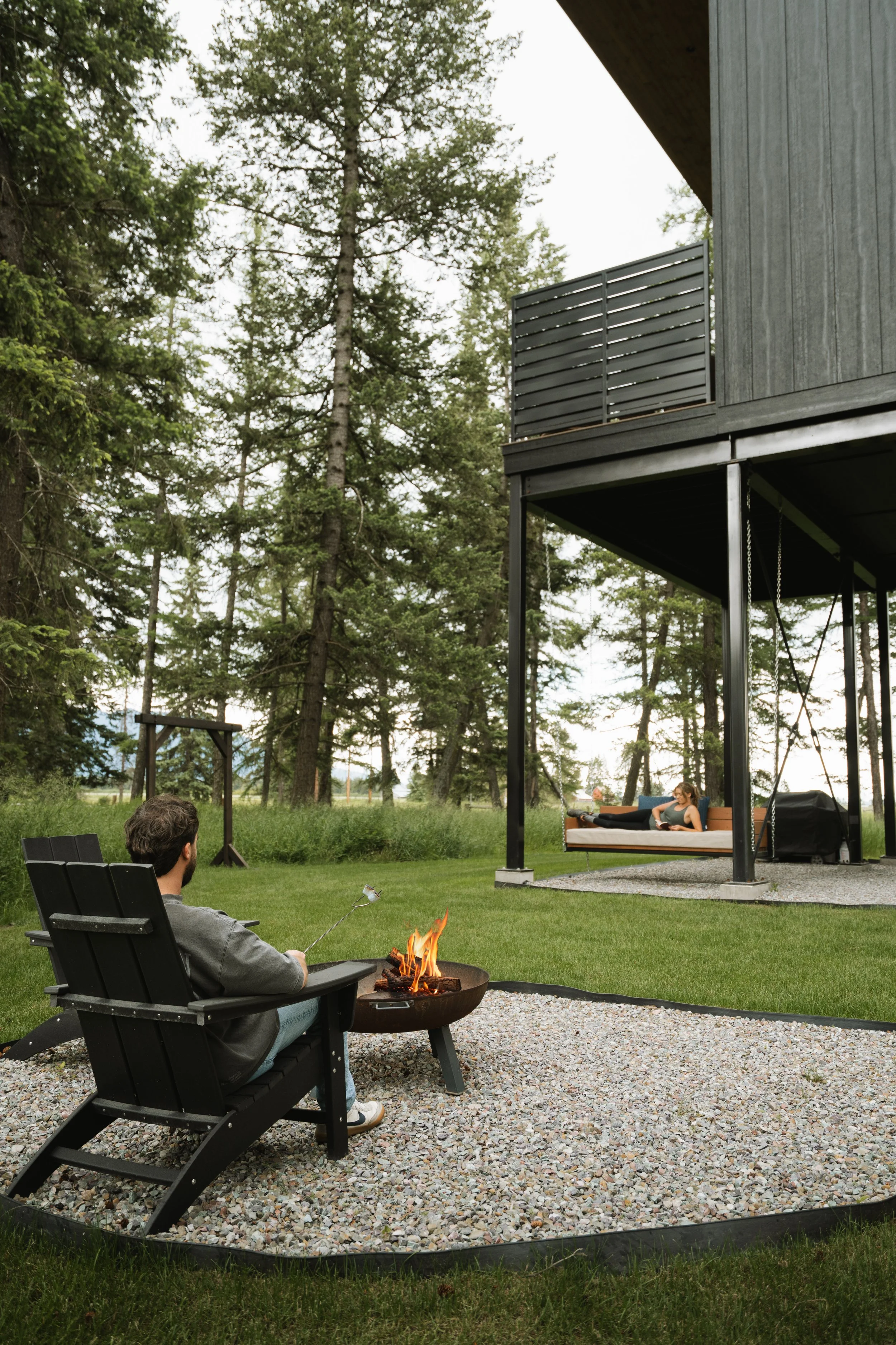 A man sitting in a black outdoor chair near a small fire pit, with a woman relaxing on a swing bed on a porch in a wooded area.