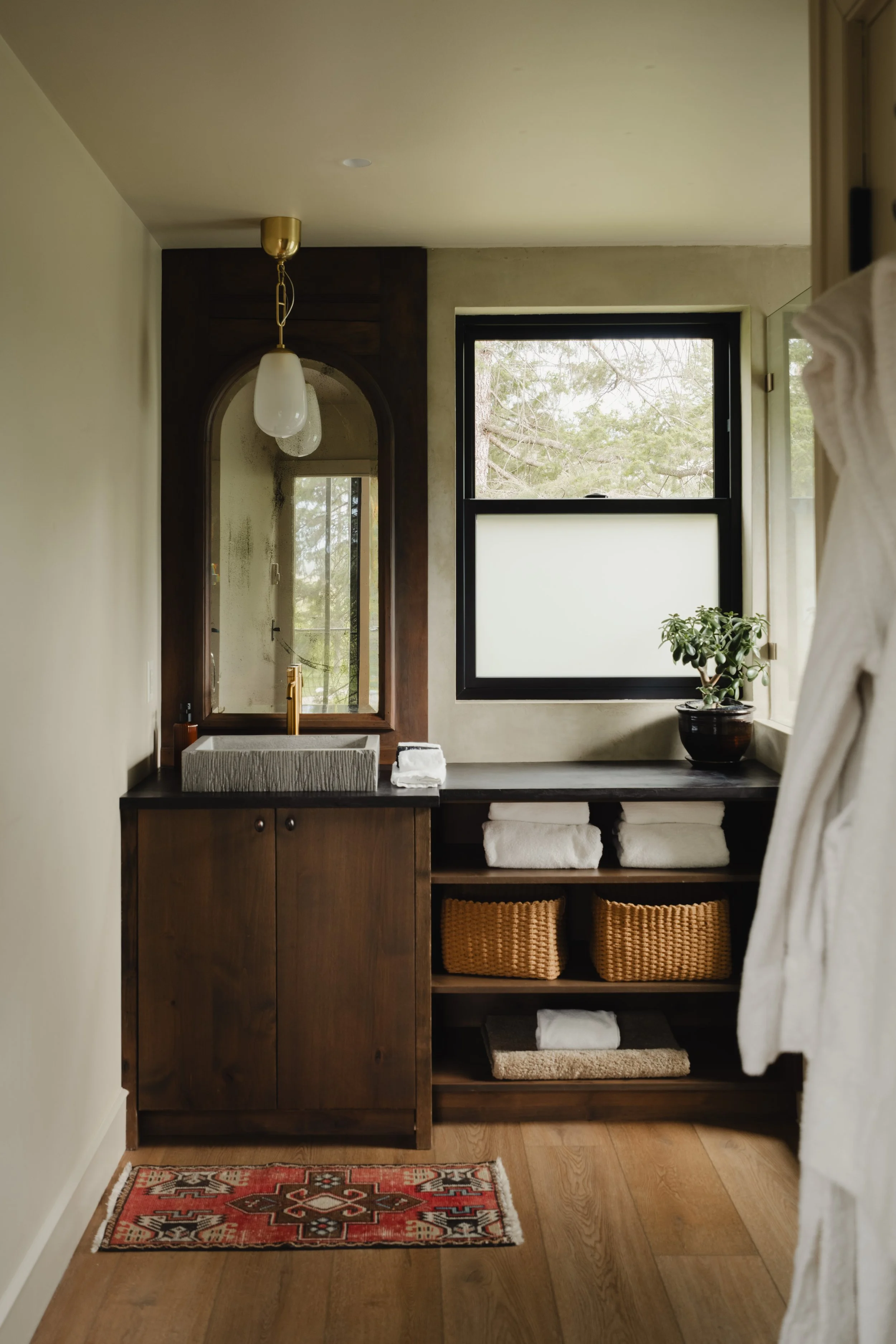 A bathroom vanity with a rectangular countertop sink, wooden cabinet, and a mirror with a curved top. There are folded towels and woven baskets on open shelves, a small plant, and a window with a view of trees.