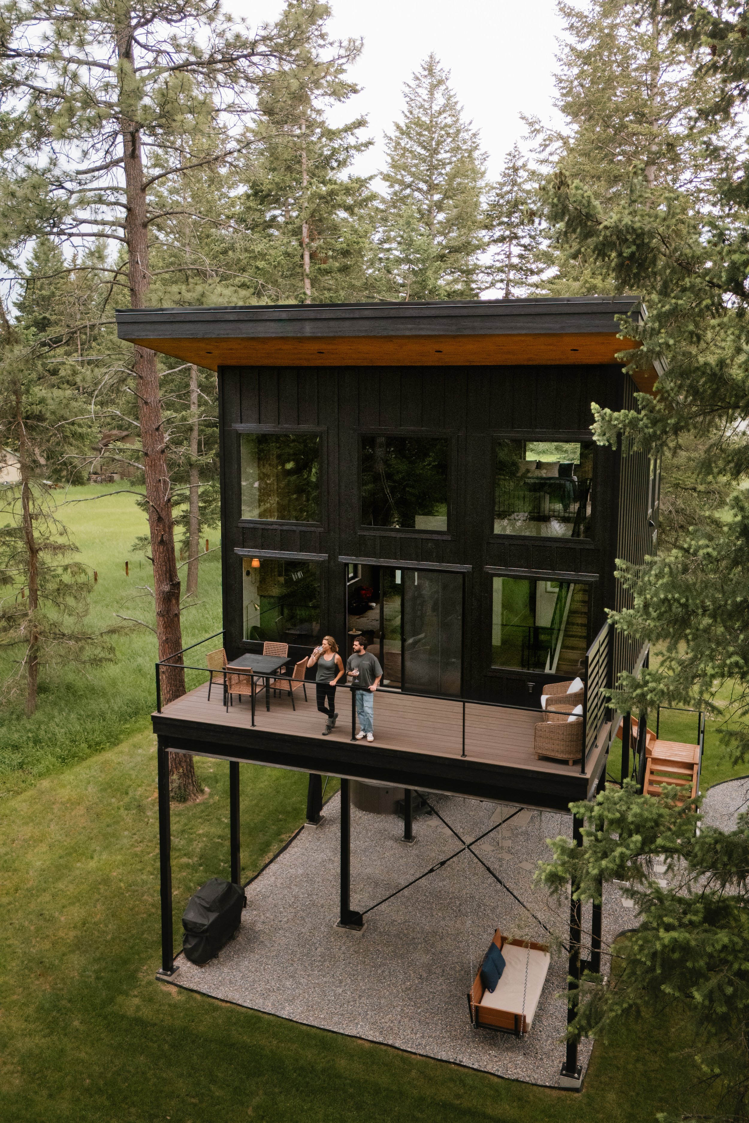 A modern black house with large windows and a wooden roof, situated on stilts among tall evergreen trees. There is a balcony with outdoor furniture, and two people are standing on it, talking. The house is surrounded by a green lawn and a gravel pati