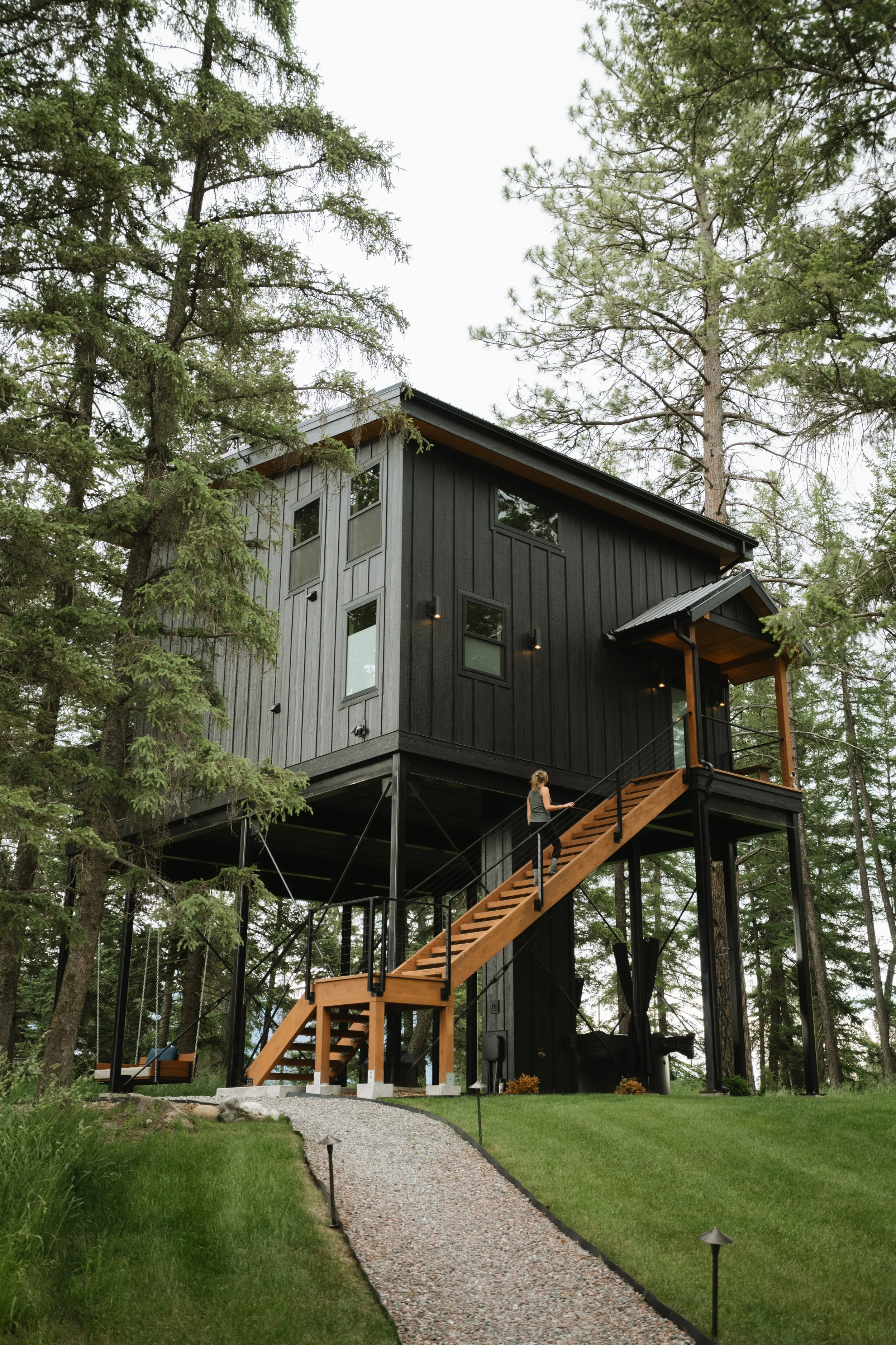 A black modern house with large windows is elevated on stilts among tall pine trees. A wooden staircase leads up to a small balcony in the front, and there is a gravel pathway lined with small outdoor lights in the surrounding yard.