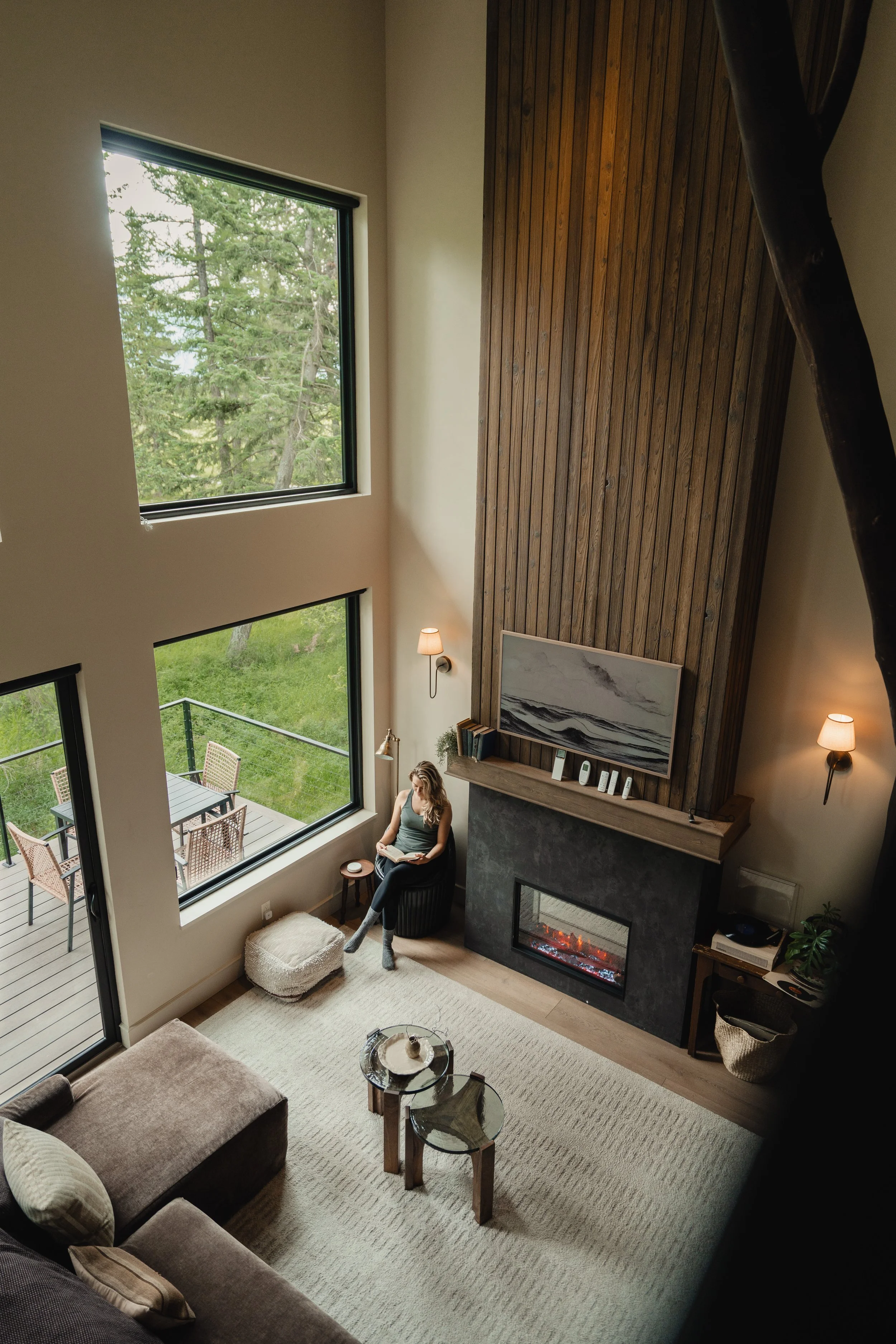 Living room with a woman sitting by a large window, reading a book, next to a fireplace with a wooden mantel and artwork on the wall. There is a patio with outdoor furniture visible through the window.