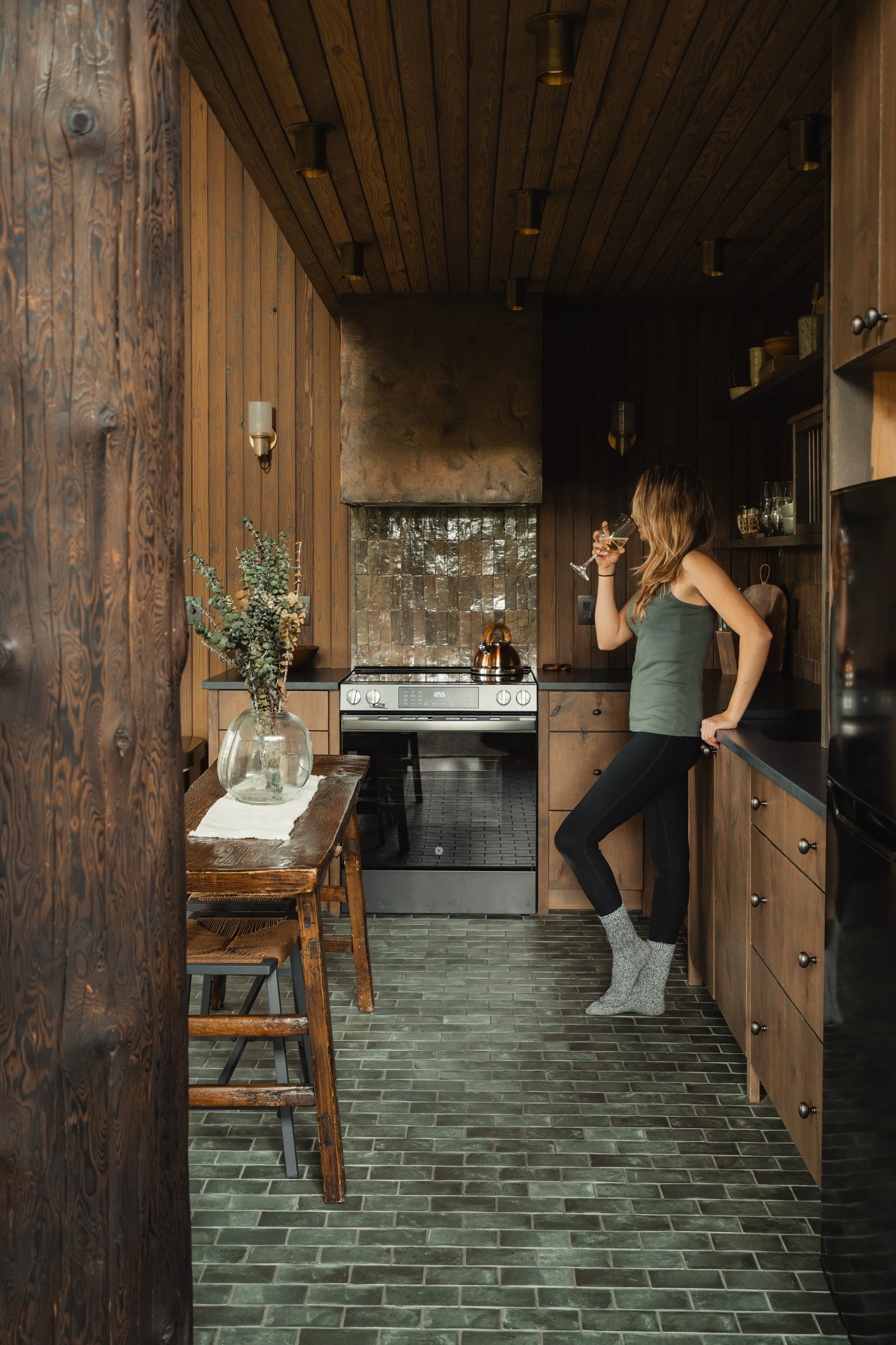 A woman standing in a rustic kitchen drinking from a wine glass, wearing a green tank top, black leggings, and gray socks, with wood panel walls, green tile floor, and wooden cabinetry.