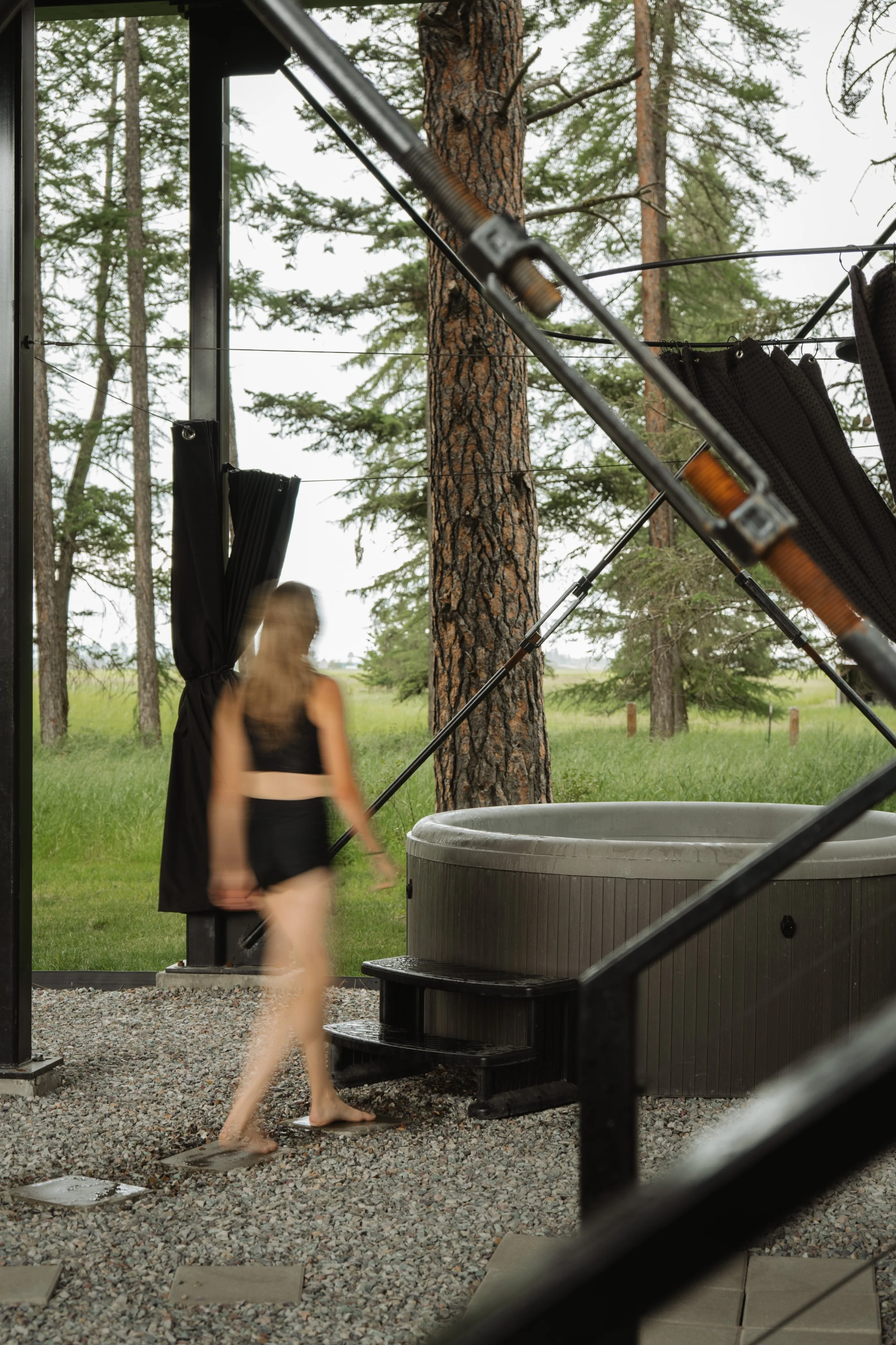 A woman in black swimwear walking on a gravel pathway outside near a hot tub, with trees and a cloudy sky in the background. The image is taken from inside a structure, looking out.