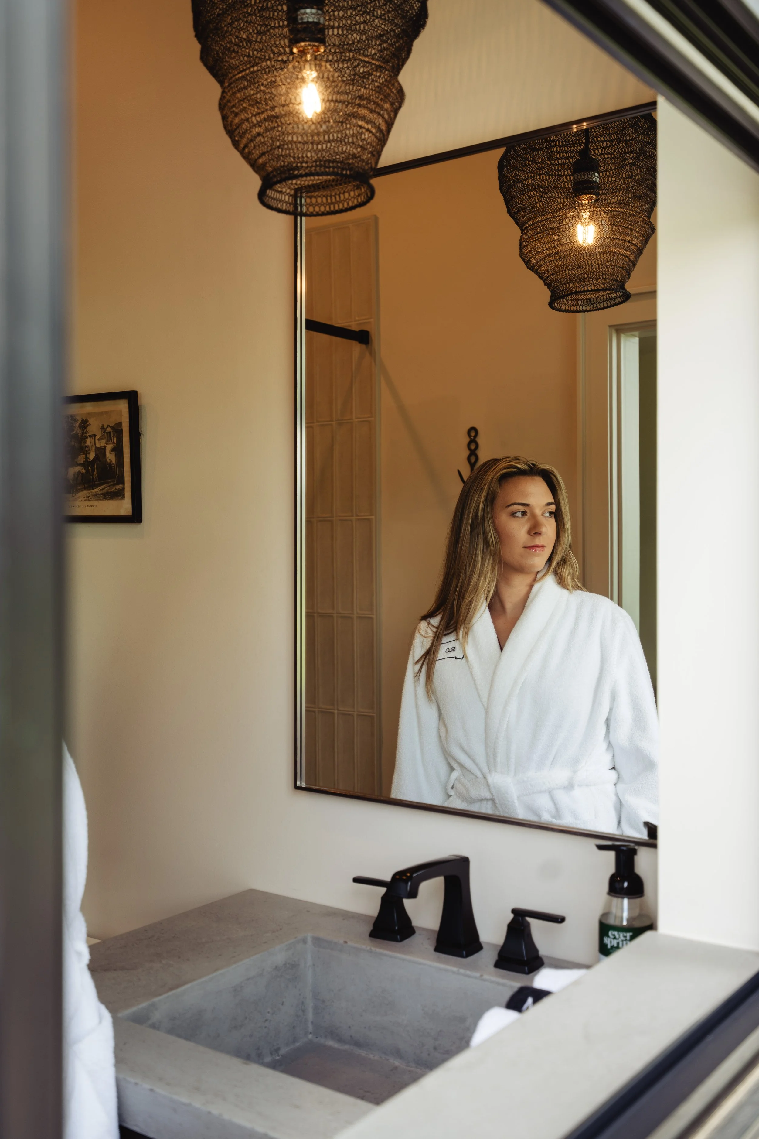 A woman in a white bathrobe looking into a mirror in a bathroom. The bathroom features a stone sink, black faucet, and wall-mounted light fixtures with woven shades.