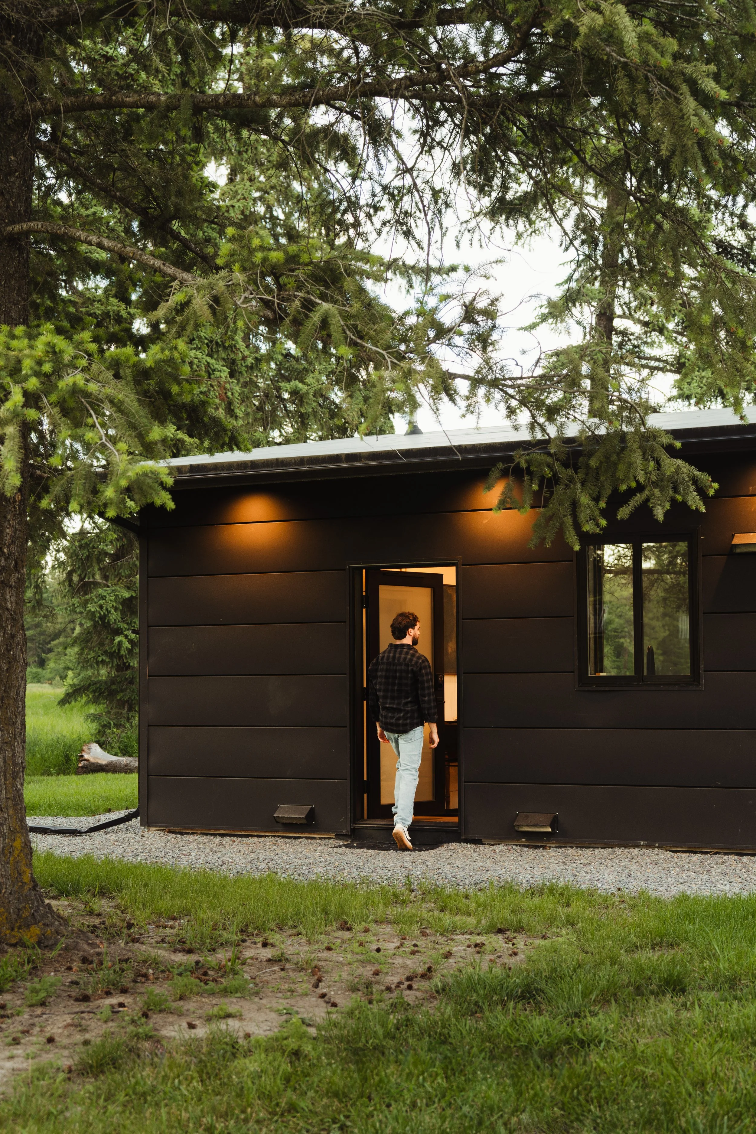 A person with curly hair wearing a plaid shirt and light jeans walking into a black modern tiny house with an open door, surrounded by green grass, trees, and a gravel path.