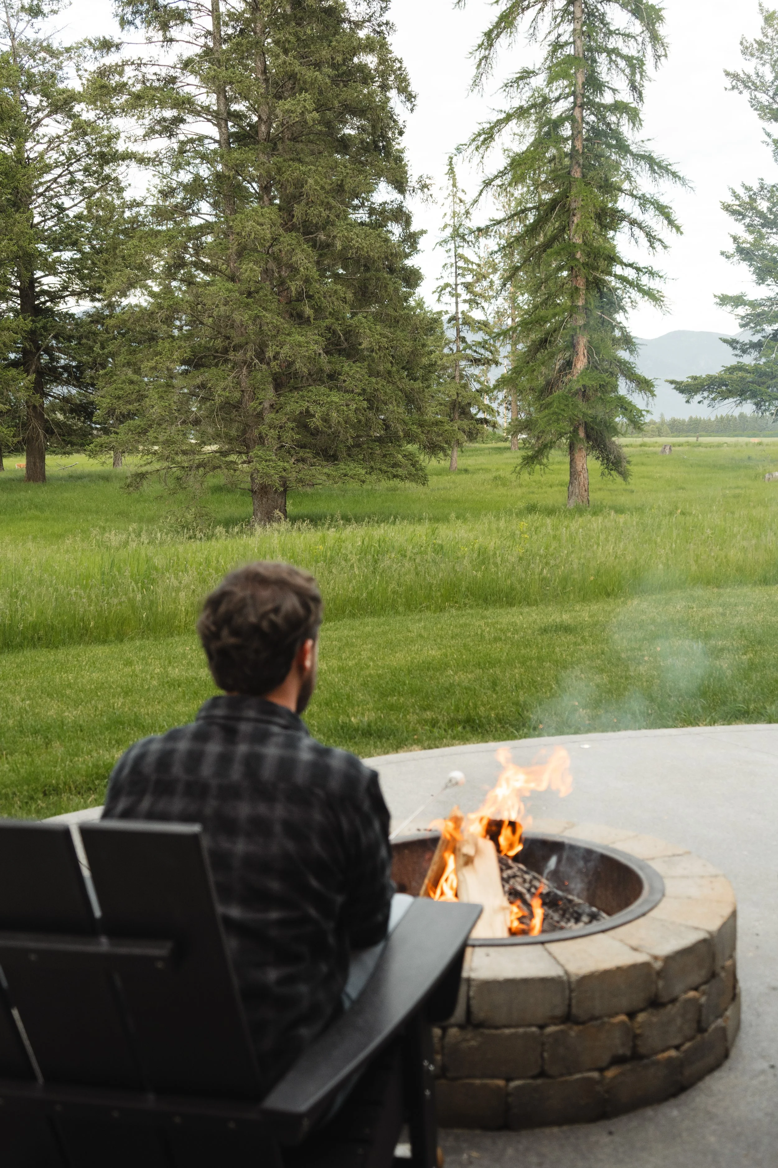 A person sitting in a black chair by a fire pit outdoors, overlooking a grassy field with trees and mountains in the background.