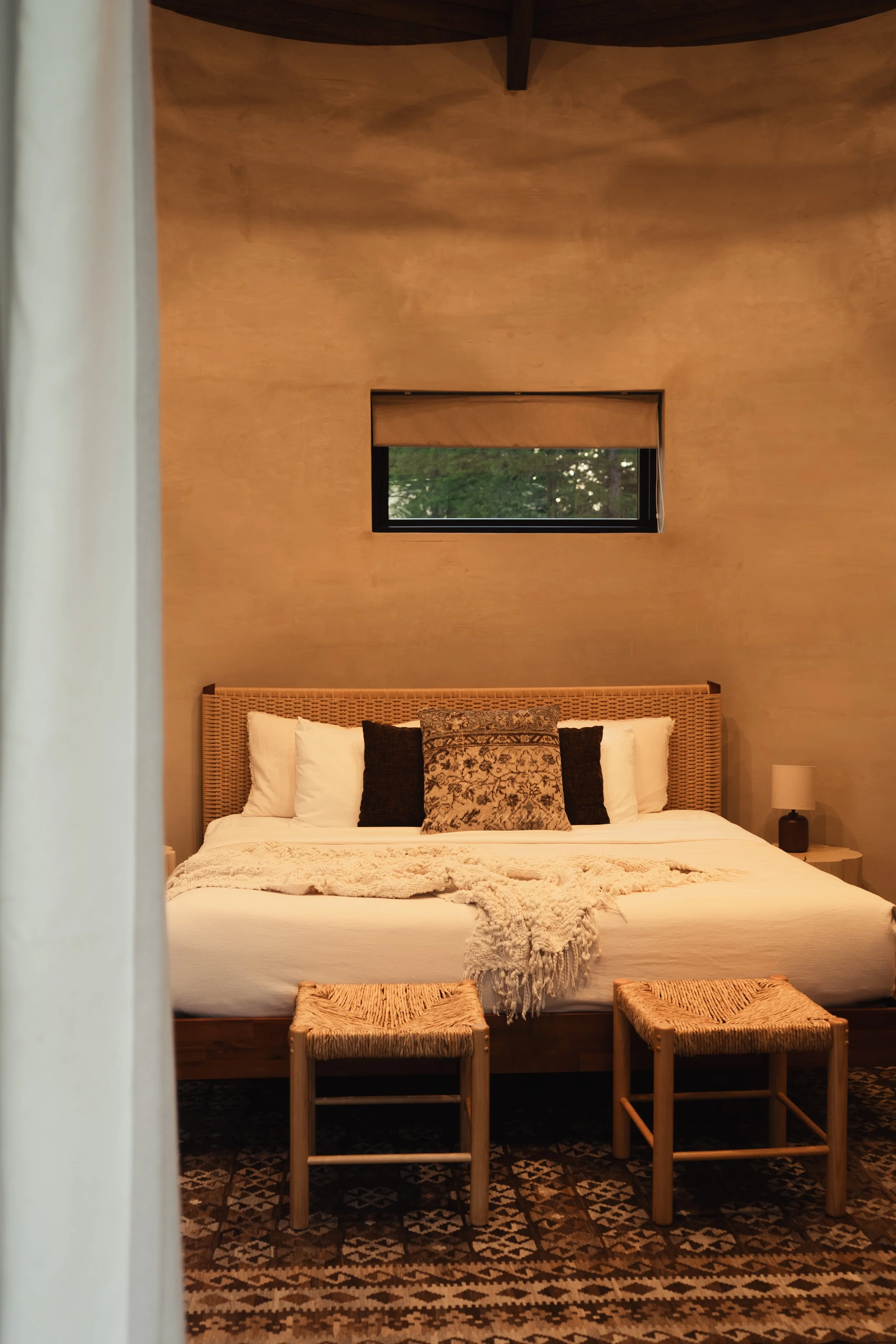 A bedroom with a bed, decorative pillows, three small rattan stools, a nightstand with a lamp, a window showing trees outside, and warm-colored textured walls.