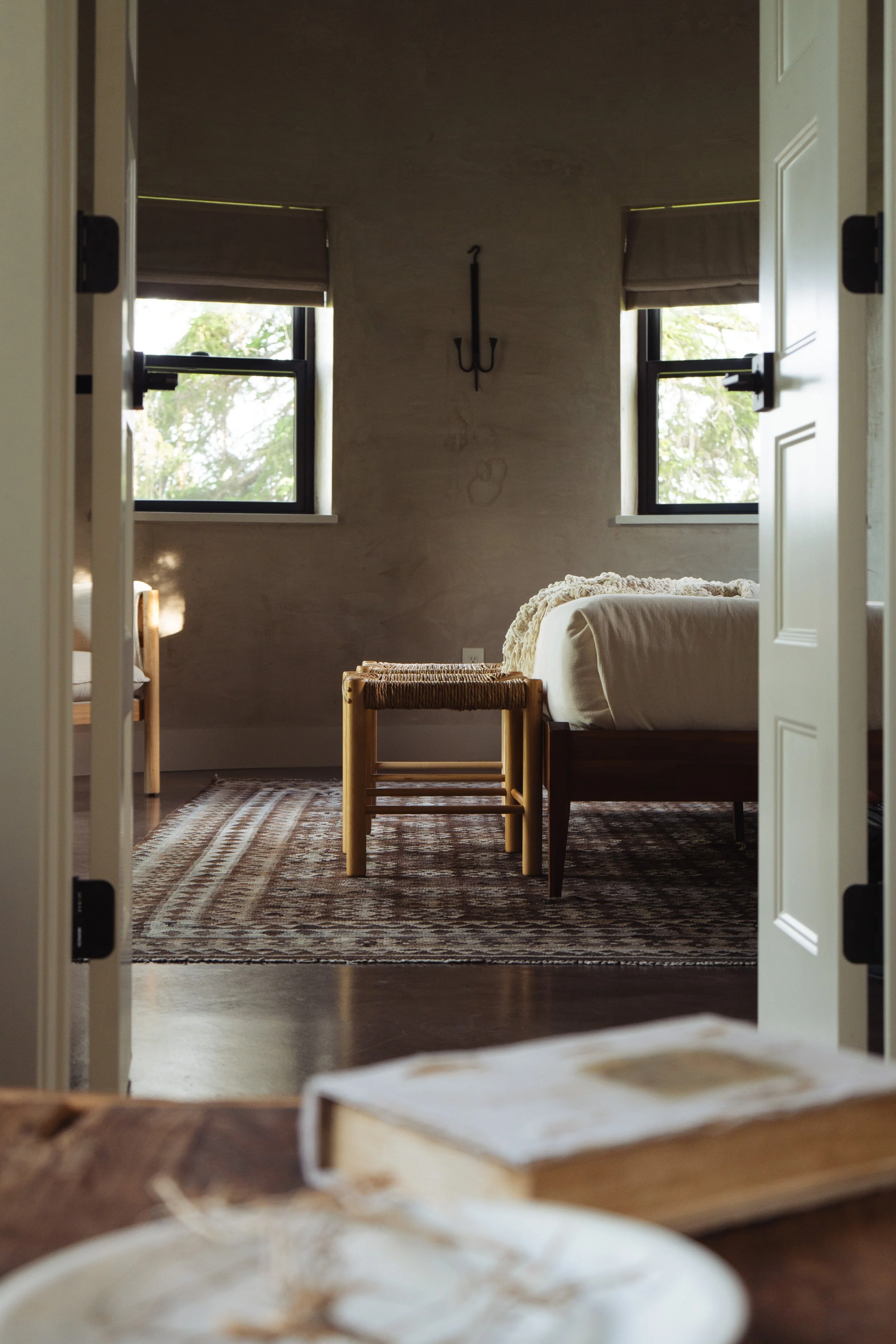 Room with two open windows, a wooden bed, a small stool, a patterned rug, and a wall sconce, viewed through a doorway.