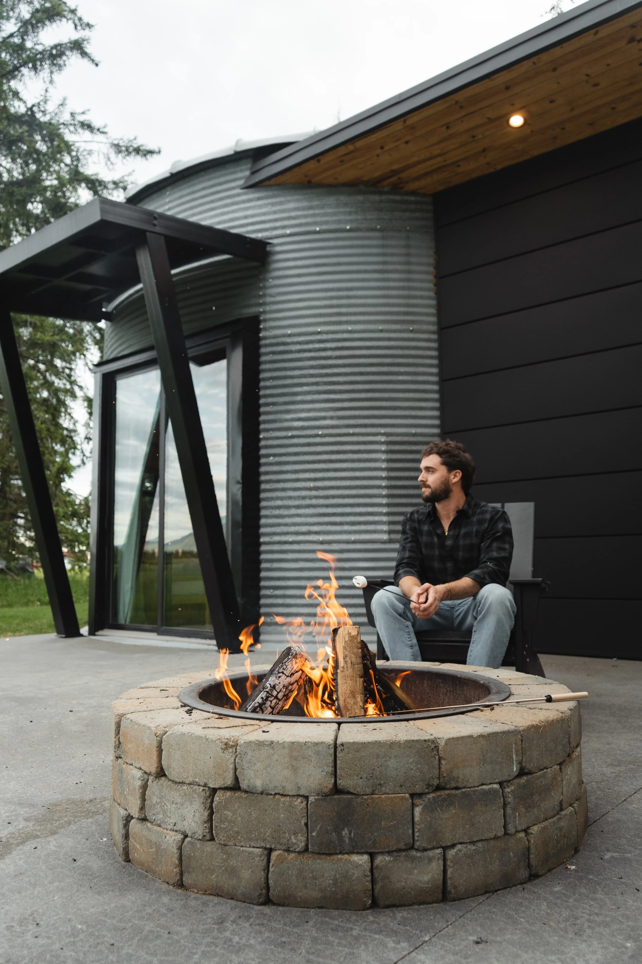 A man sitting outside by a fire pit on a patio in front of a modern house with black and corrugated metal exterior, a glass door, and a wooden roof