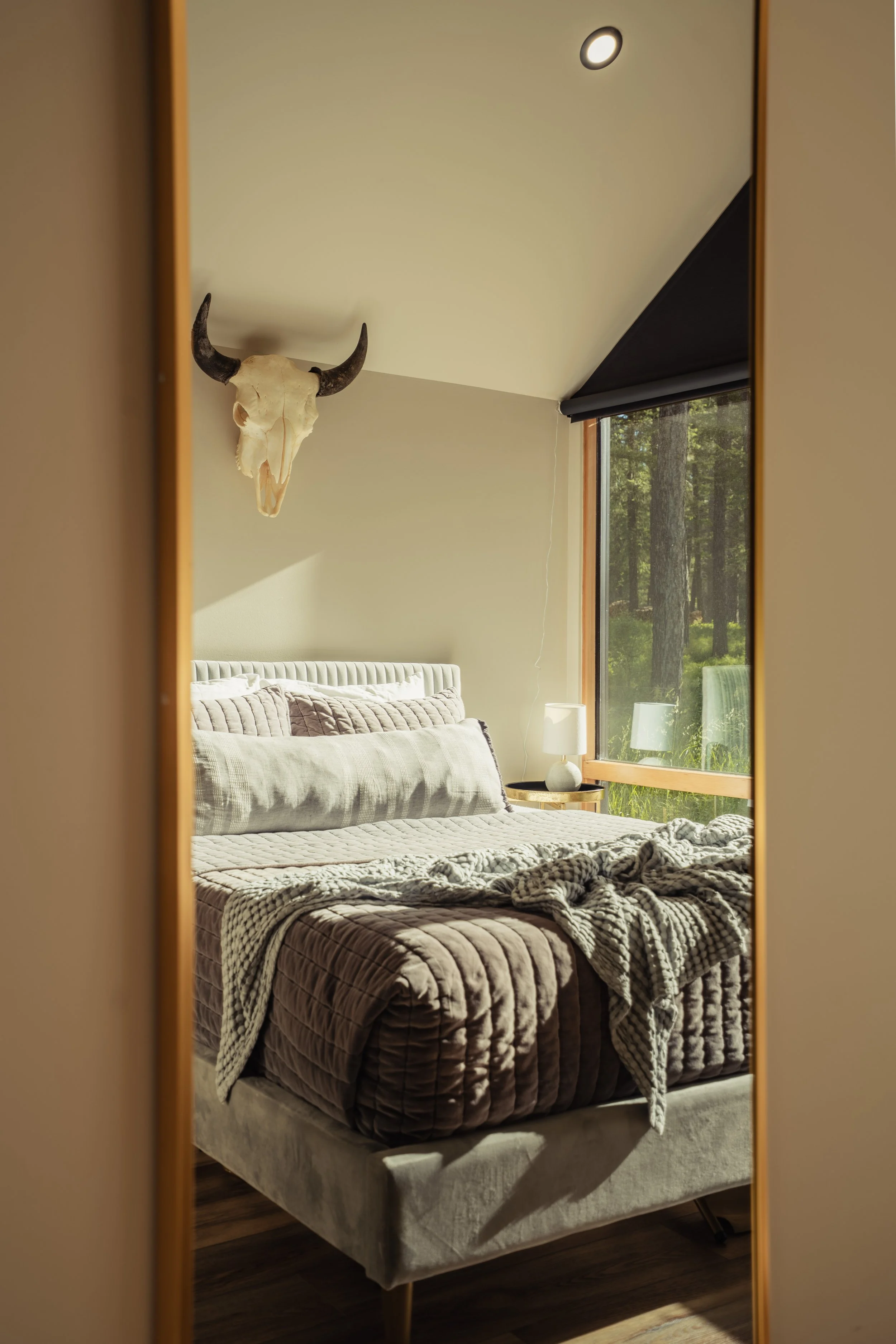 A bedroom with a bed covered in a quilted blanket, a nightstand with a lamp, a window with a view of trees, and a skull with horns mounted on the wall.