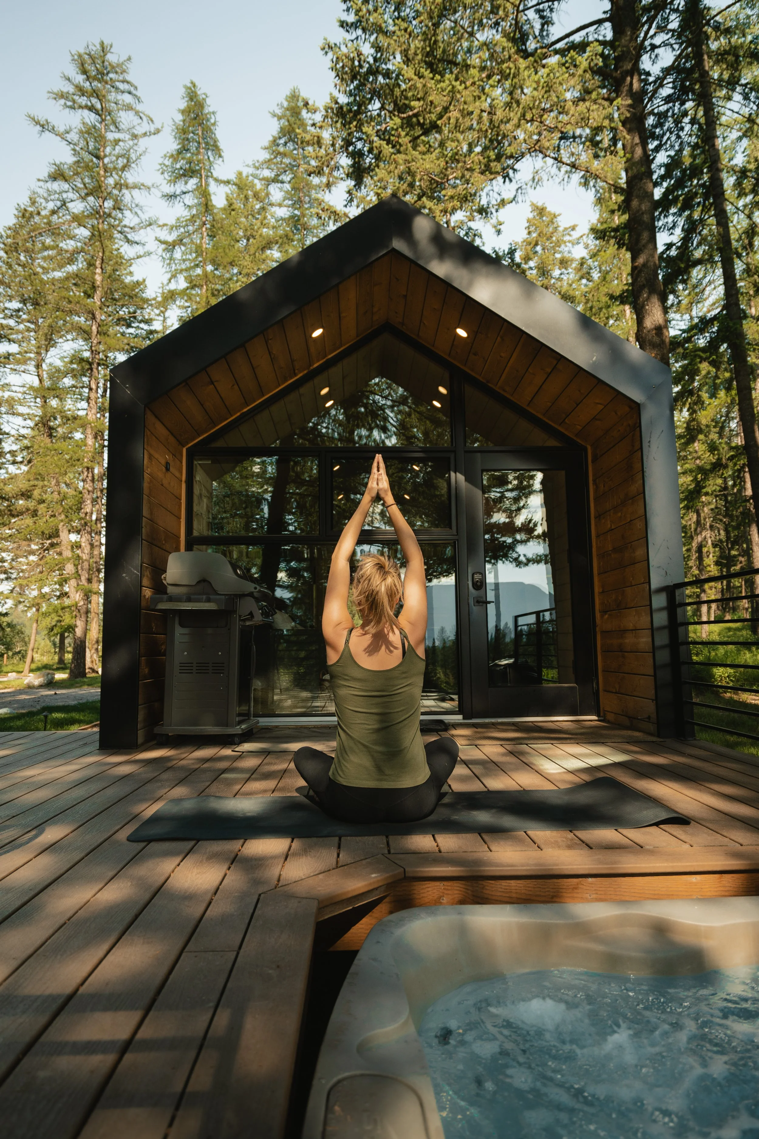 A woman sits cross-legged on a yoga mat on a wooden deck outside a modern cabin, practicing yoga with arms raised above her head in a prayer position. The background features tall pine trees and a clear sky, with a hot tub visible in the foreground.