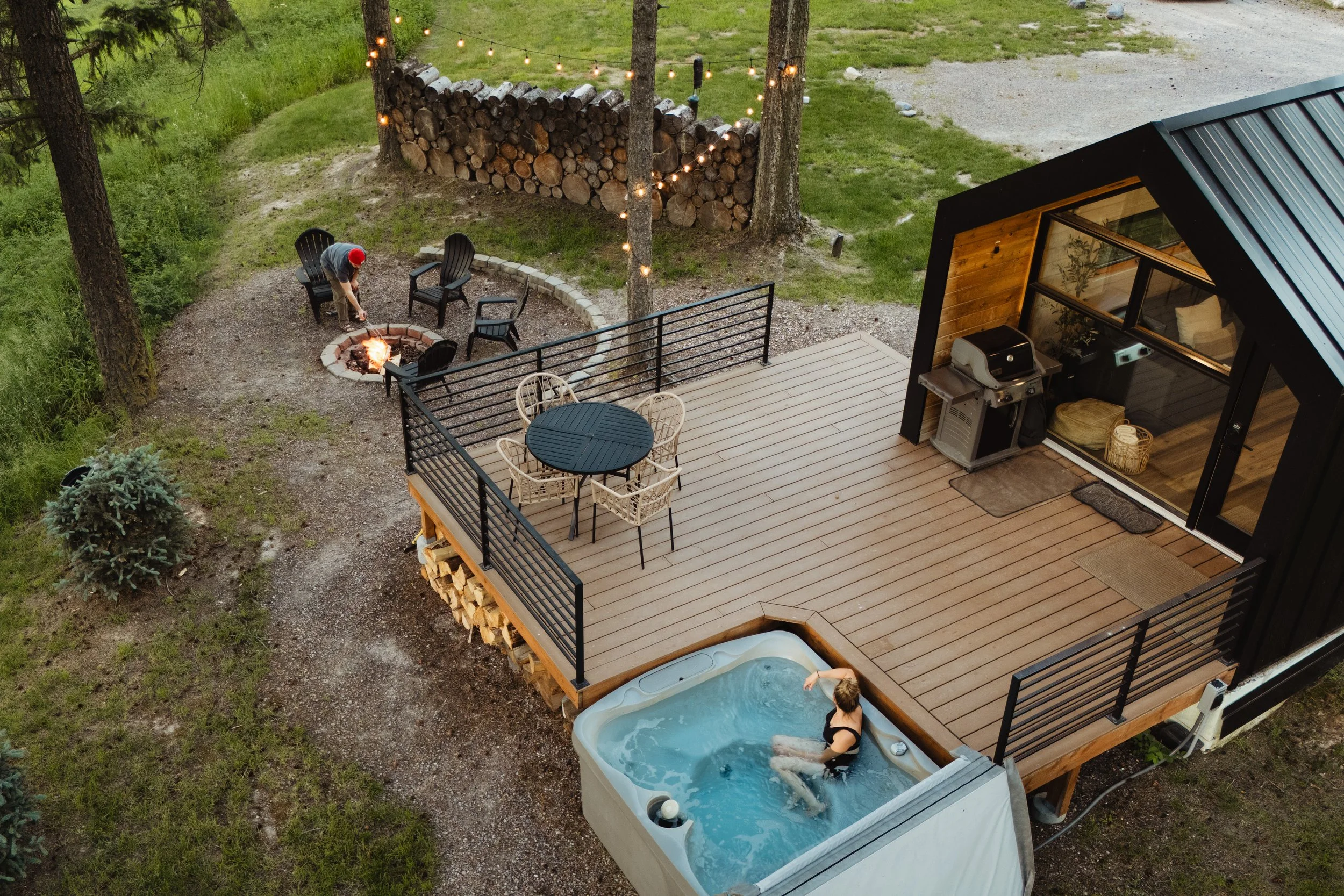 A backyard patio with a fire pit, outdoor seating, and a hot tub. There is a person in the hot tub and a person near the fire pit. A stack of firewood and string lights are visible, along with a modern house with a deck.