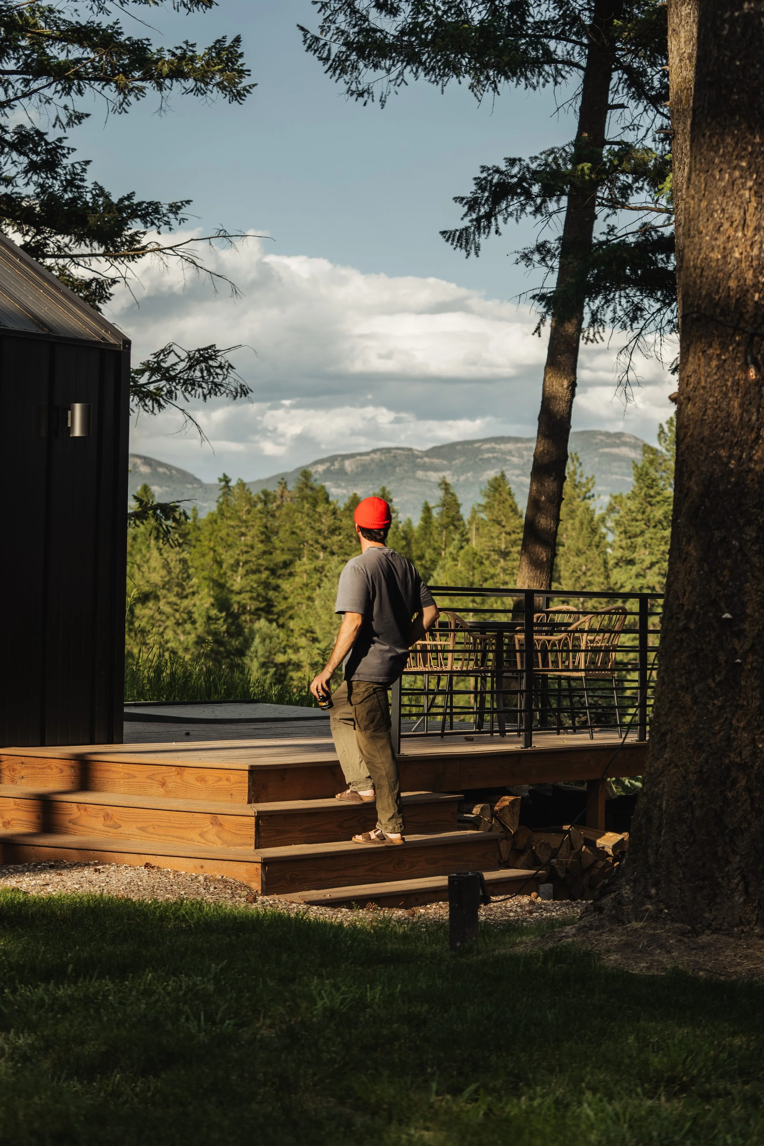 A person with a red hat and gray shirt walking down wooden stairs on a deck outdoors, with trees, mountains, and cloudy sky in the background.