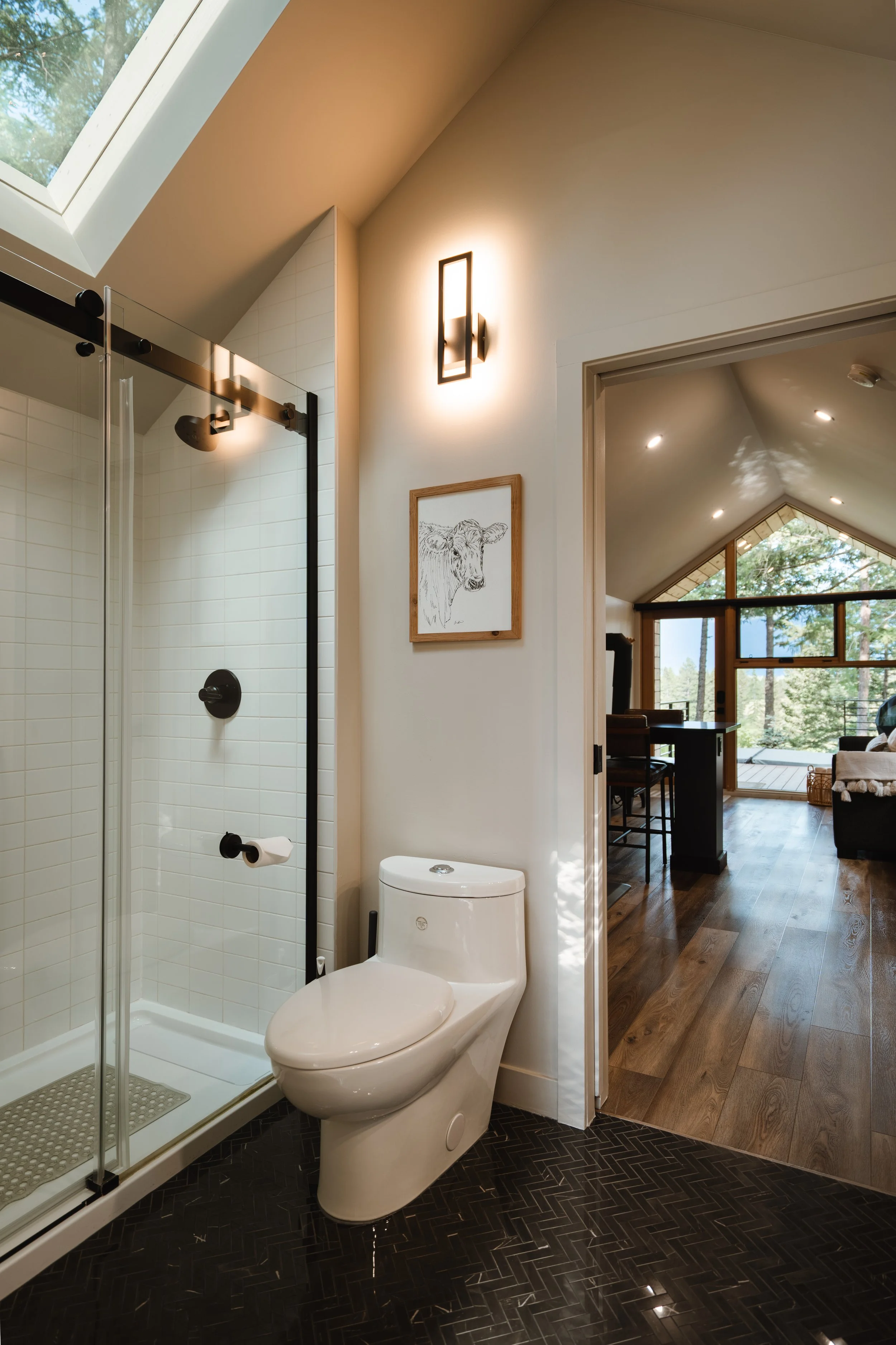 A modern bathroom with a shower and a toilet, a framed cow illustration on the wall, and view into a living space with large windows and wood floors.